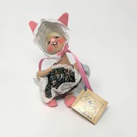 Tan felt bunny wearing a dark floral dress with white eyelet trim and a matching white bonnet with pink ribbon ties. She has pink-lined ears and wears tiny pink felt slippers. A toothy, joyful hand-painted grin and a vintage Annalee tag are visible.