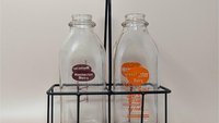 Two vintage clear glass milk bottles with red and orange Rochester Dairy logos sitting inside a black metal wire carrying crate with a wooden handle.