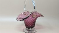 A vibrant cranberry pink glass basket with a clear arched handle. The body features a raised hobnail texture and a ruffled, flared rim, supported by a clear, scalloped pedestal base.