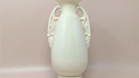 A studio photo of a tall, cream-colored ceramic vase with a glossy finish. The vase features an elegant urn shape with two ornate, scroll-style handles on the sides. It has a flared rim and sits on a tiered circular base with a beaded detail around the bottom.