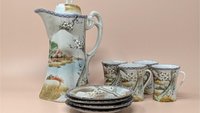 A hand-painted porcelain tea set featuring a tall teapot, four cups, and four saucers. The set depicts a traditional Japanese landscape with mountains, water, and flowering trees in soft earth tones and blue borders.