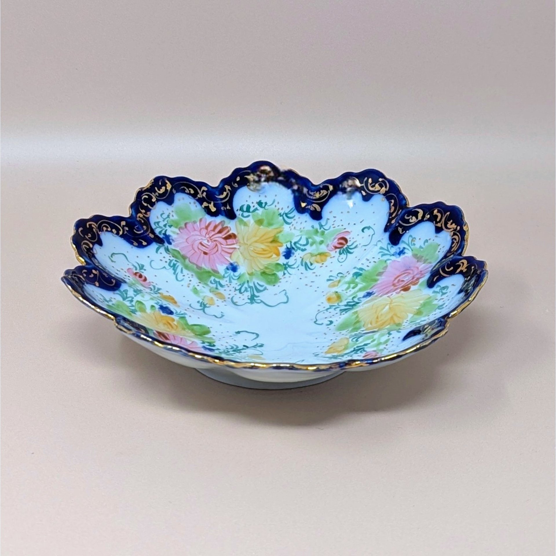 Scalloped porcelain bowl with a deep cobalt blue rim and intricate gold filigree. The interior features hand-painted pink and yellow floral bouquets with delicate green vines on a white ground.