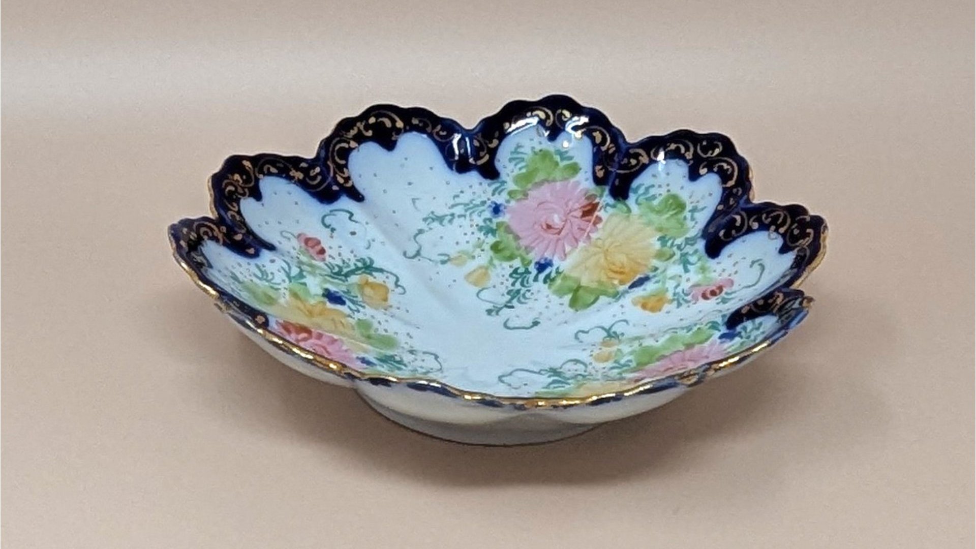 Scalloped porcelain bowl with a deep cobalt blue rim and intricate gold filigree. The interior features hand-painted pink and yellow floral bouquets with delicate green vines on a white ground.