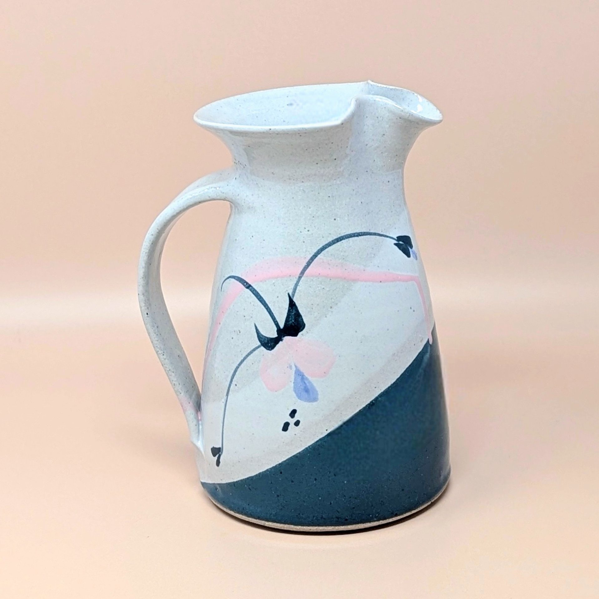 Speckled cream earthenware pitcher with a wide flared rim and sturdy handle. Features a hand-painted abstract floral design in teal, pink, and indigo with a diagonal teal color-block base.