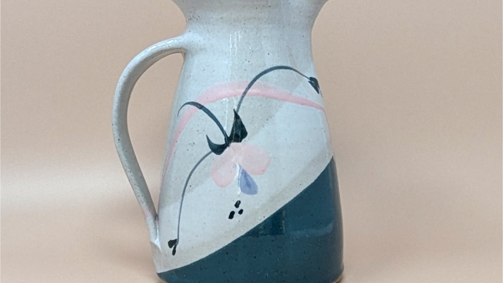 Speckled cream earthenware pitcher with a wide flared rim and sturdy handle. Features a hand-painted abstract floral design in teal, pink, and indigo with a diagonal teal color-block base.