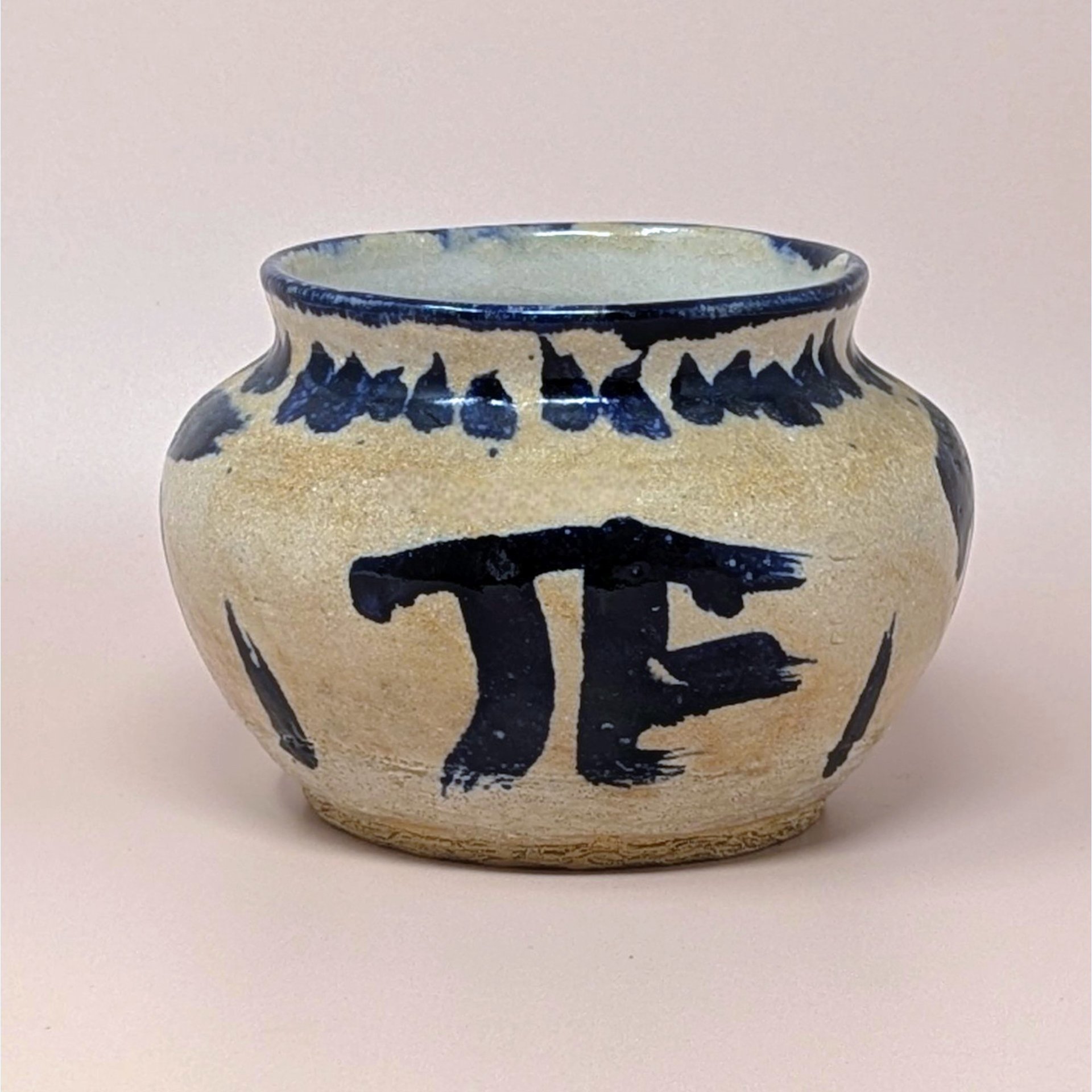 Hand-thrown pottery bowl with a globular shape and a sandy, textured tan glaze. Featuring bold, gestural cobalt blue brushstrokes including a large "PI" or "TF" symbol and a saw-tooth rim pattern.