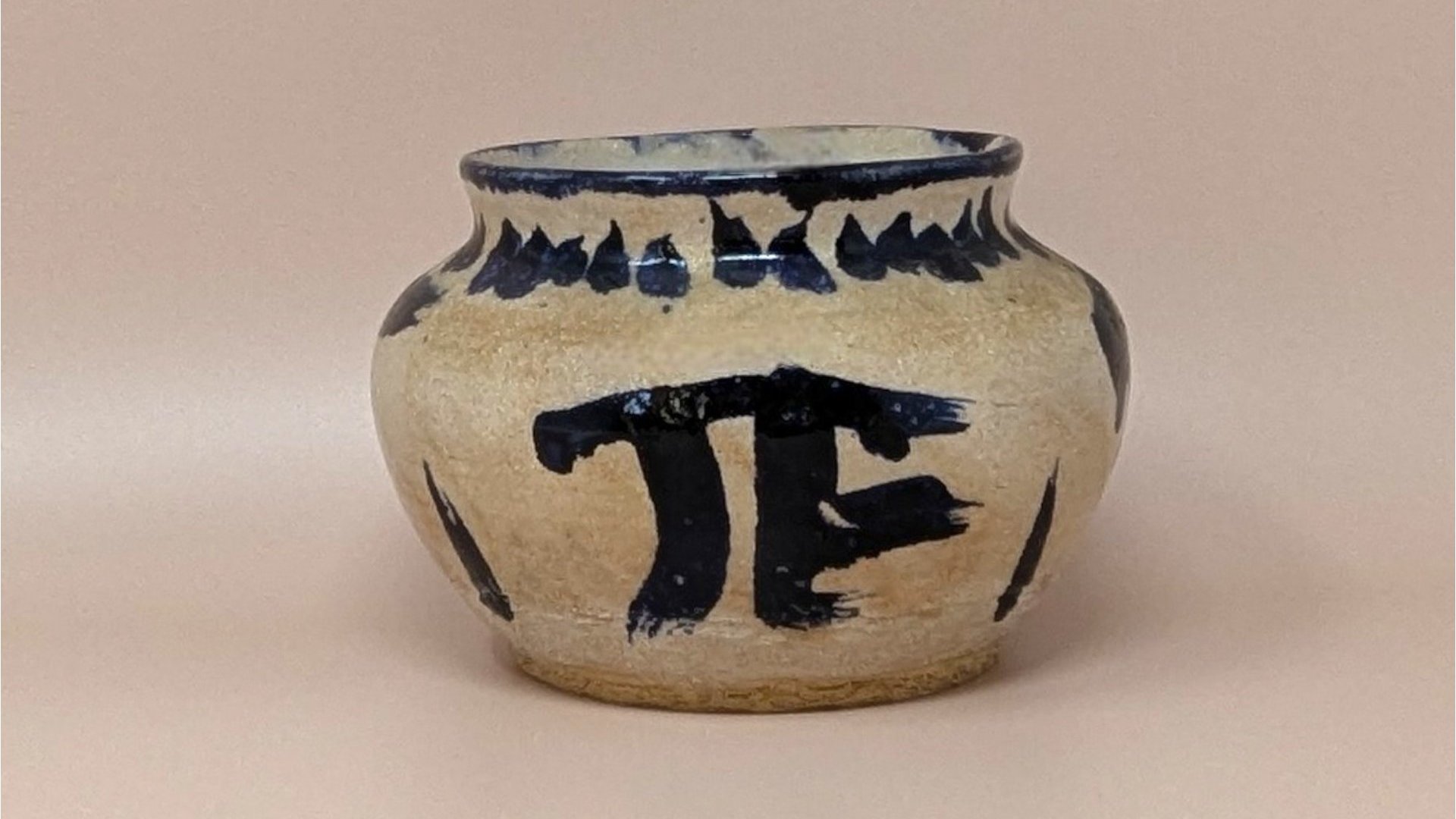 Hand-thrown pottery bowl with a globular shape and a sandy, textured tan glaze. Featuring bold, gestural cobalt blue brushstrokes including a large "PI" or "TF" symbol and a saw-tooth rim pattern.