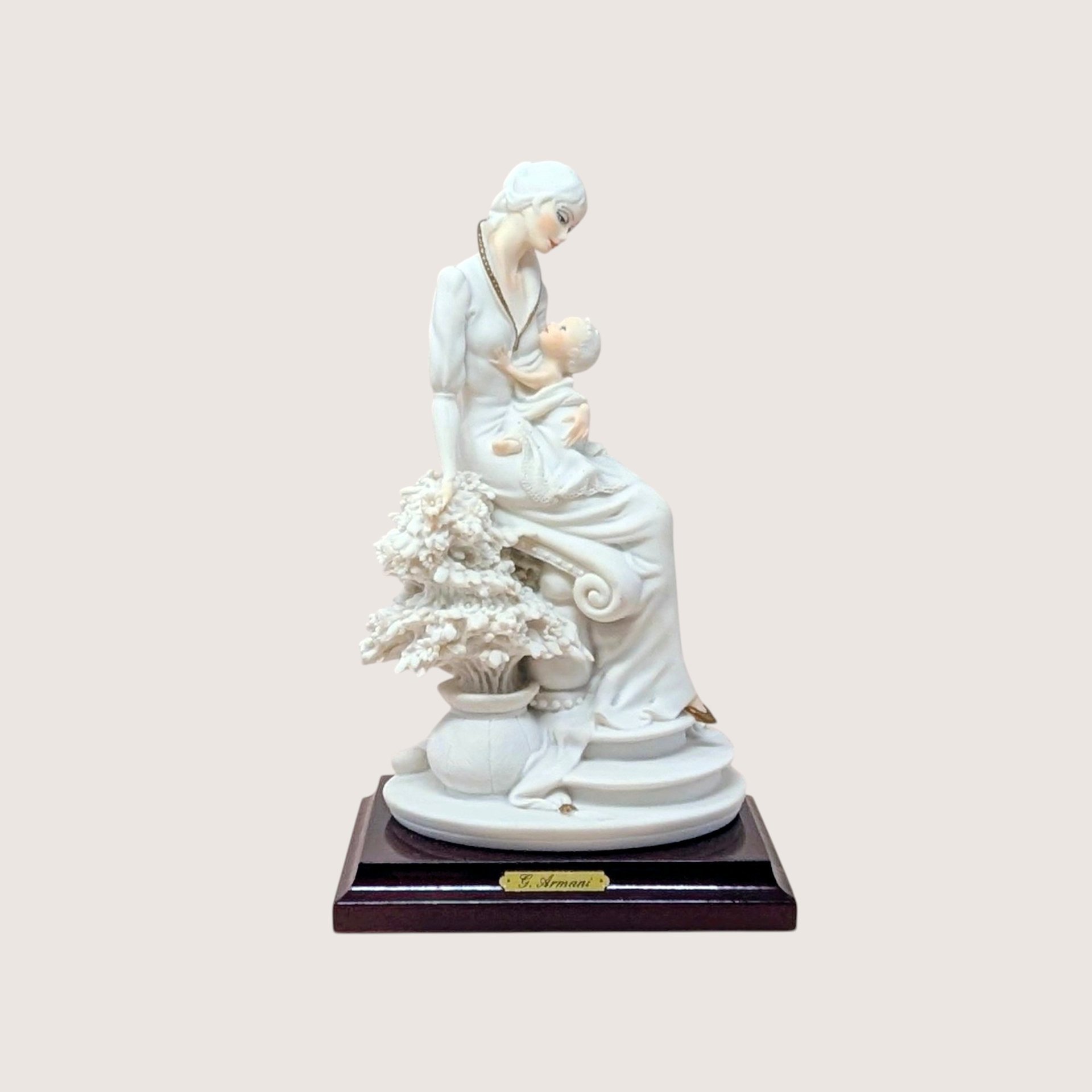 Giuseppe Armani "Maternal" porcelain figurine of a mother cradling an infant, seated beside a flowering tree. Set on a dark wood square pedestal base with a gold nameplate. Matte white and cream finish.