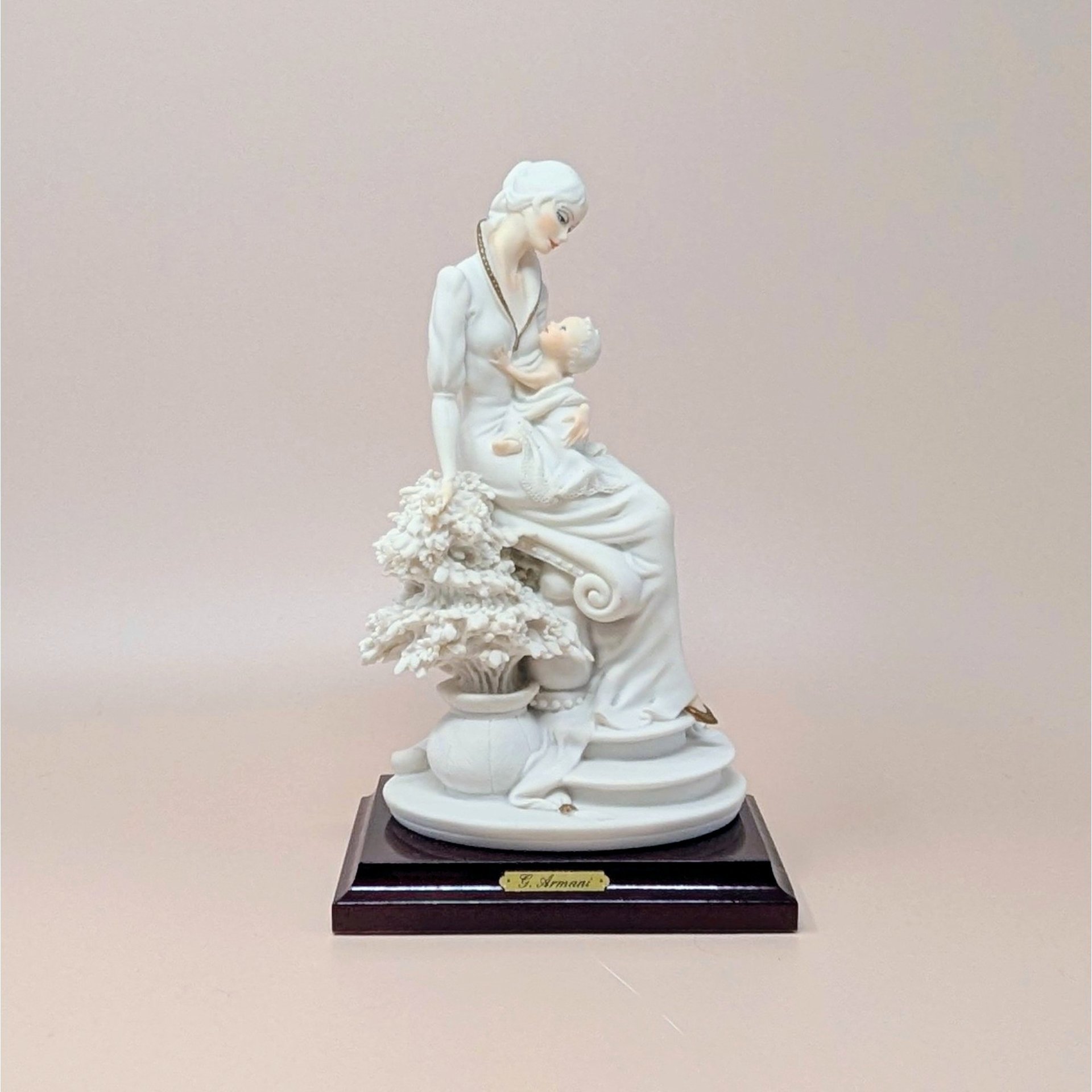 Giuseppe Armani "Maternal" porcelain figurine of a mother cradling an infant, seated beside a flowering tree. Set on a dark wood square pedestal base with a gold nameplate. Matte white and cream finish.