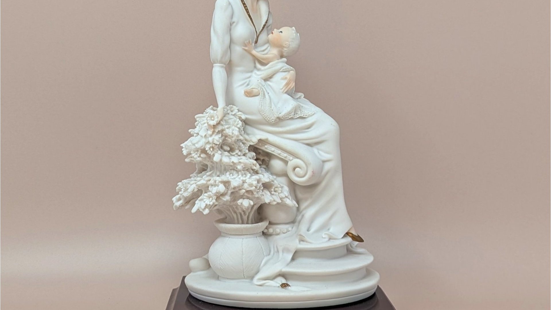 Giuseppe Armani "Maternal" porcelain figurine of a mother cradling an infant, seated beside a flowering tree. Set on a dark wood square pedestal base with a gold nameplate. Matte white and cream finish.
