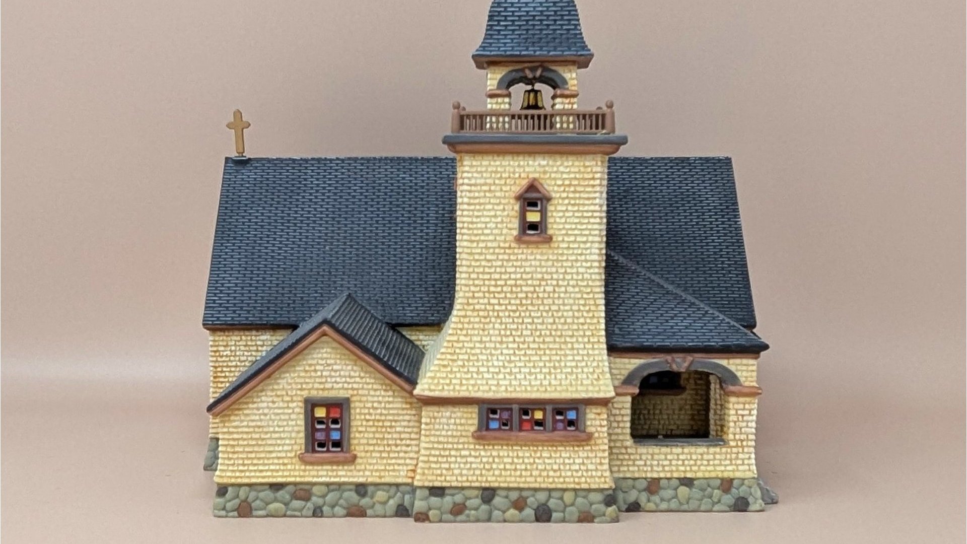 Dept. 56 Seasons Bay "Chapel on the Hill." Miniature cream-colored stone chapel with a tall dark grey steeple, stained glass windows, and a cobblestone base. Matte finish porcelain.