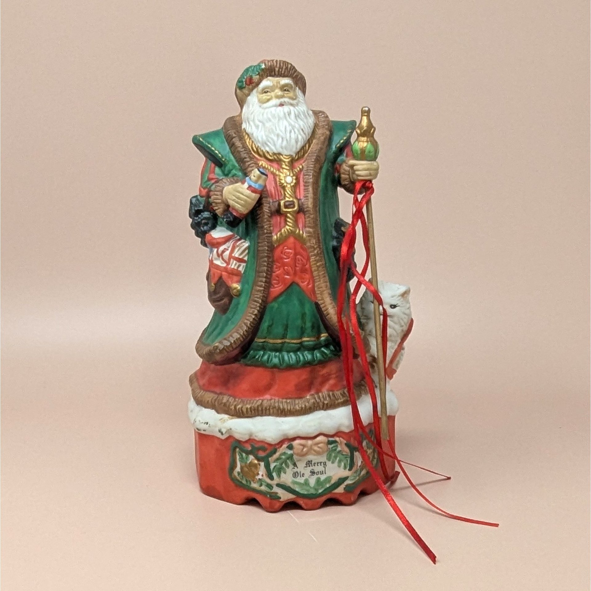A hand-painted ceramic Old World Santa figurine on a musical base. He wears a green and red robe, holding a staff with red ribbons and a nutcracker. A white bear cub sits at his foot. The red base reads "Merry Ole Soul." The style is glossy and traditional against a plain beige background.