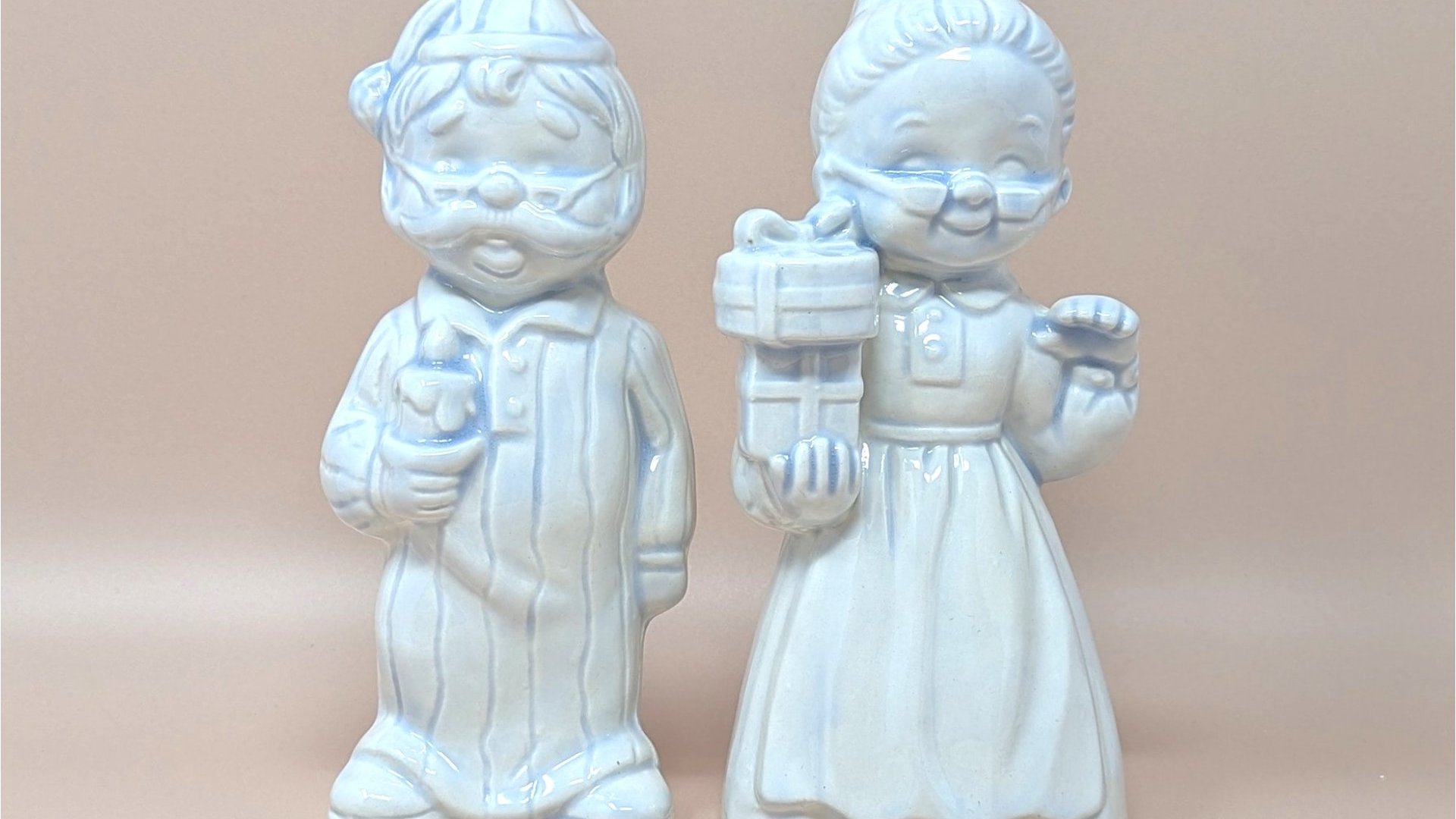 Pair of 7-inch ceramic figurines of an elderly man in striped pajamas with a candle and a woman in a nightgown holding a gift. White pearlescent luster glaze with soft blue accents.