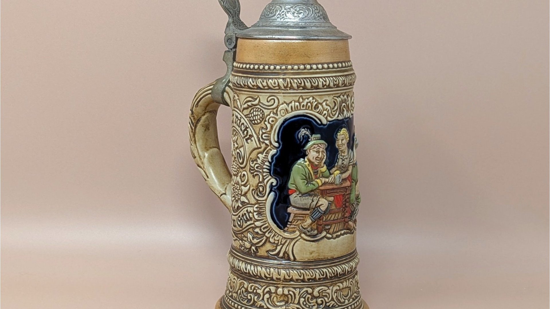 Tall Ceramarte Brazil ceramic stein with a pewter-style metal lid. High-relief tavern scene with three figures at a table against a cobalt blue background. Earthy tan and gold glaze. 10 inches tall.