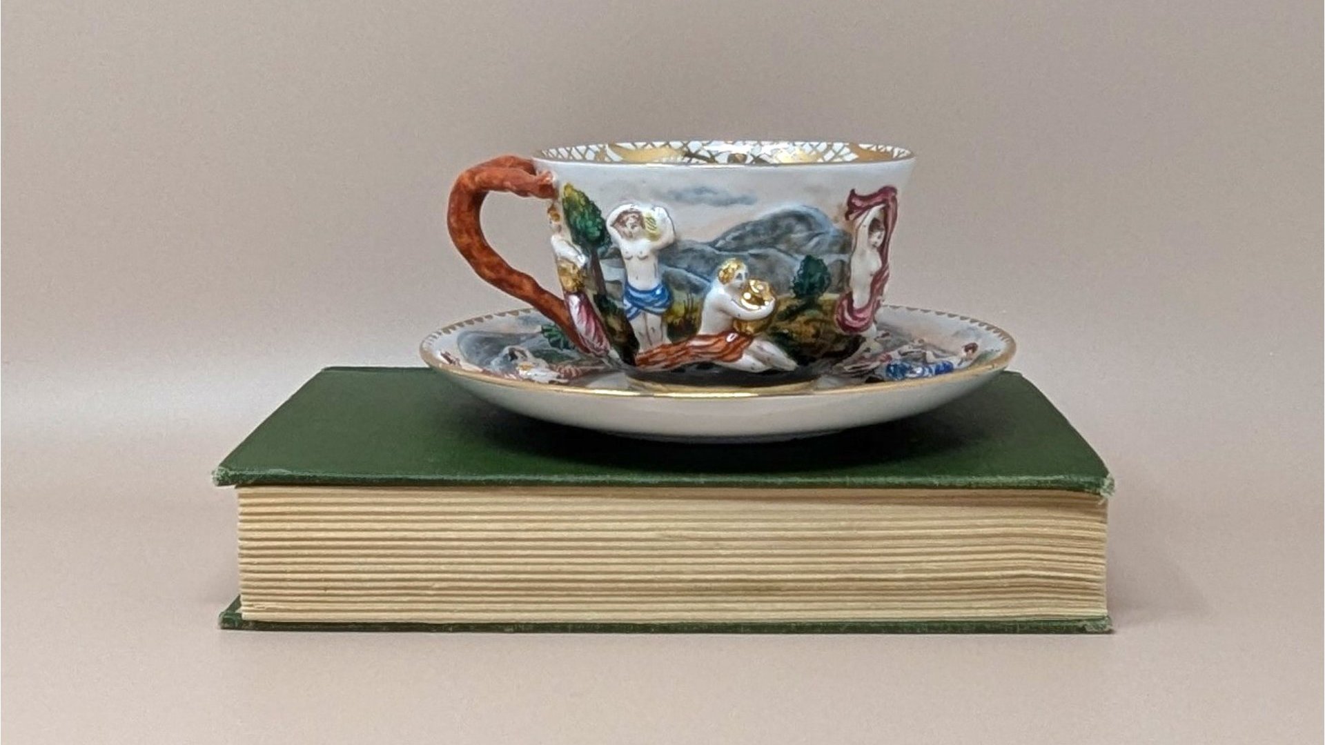 Italian Capodimonte porcelain cup and saucer set. Features high-relief, hand-painted classical figures in a woodland landscape. Intricate gold gilt interior accents and a rustic coral-style handle. Cup 2.5", Saucer 6".