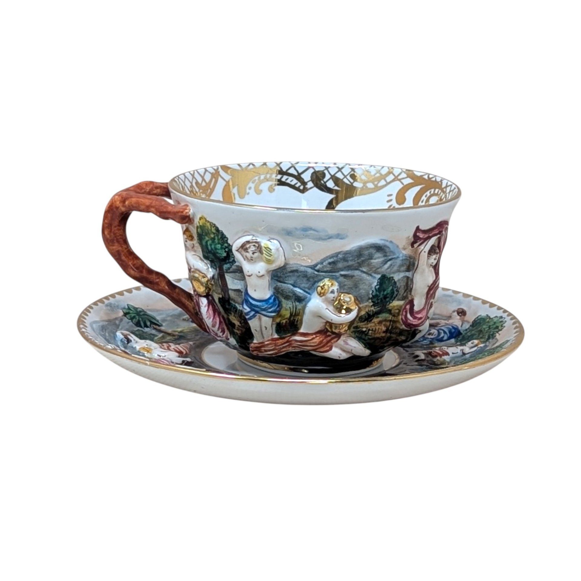 Italian Capodimonte porcelain cup and saucer set. Features high-relief, hand-painted classical figures in a woodland landscape. Intricate gold gilt interior accents and a rustic coral-style handle. Cup 2.5", Saucer 6".