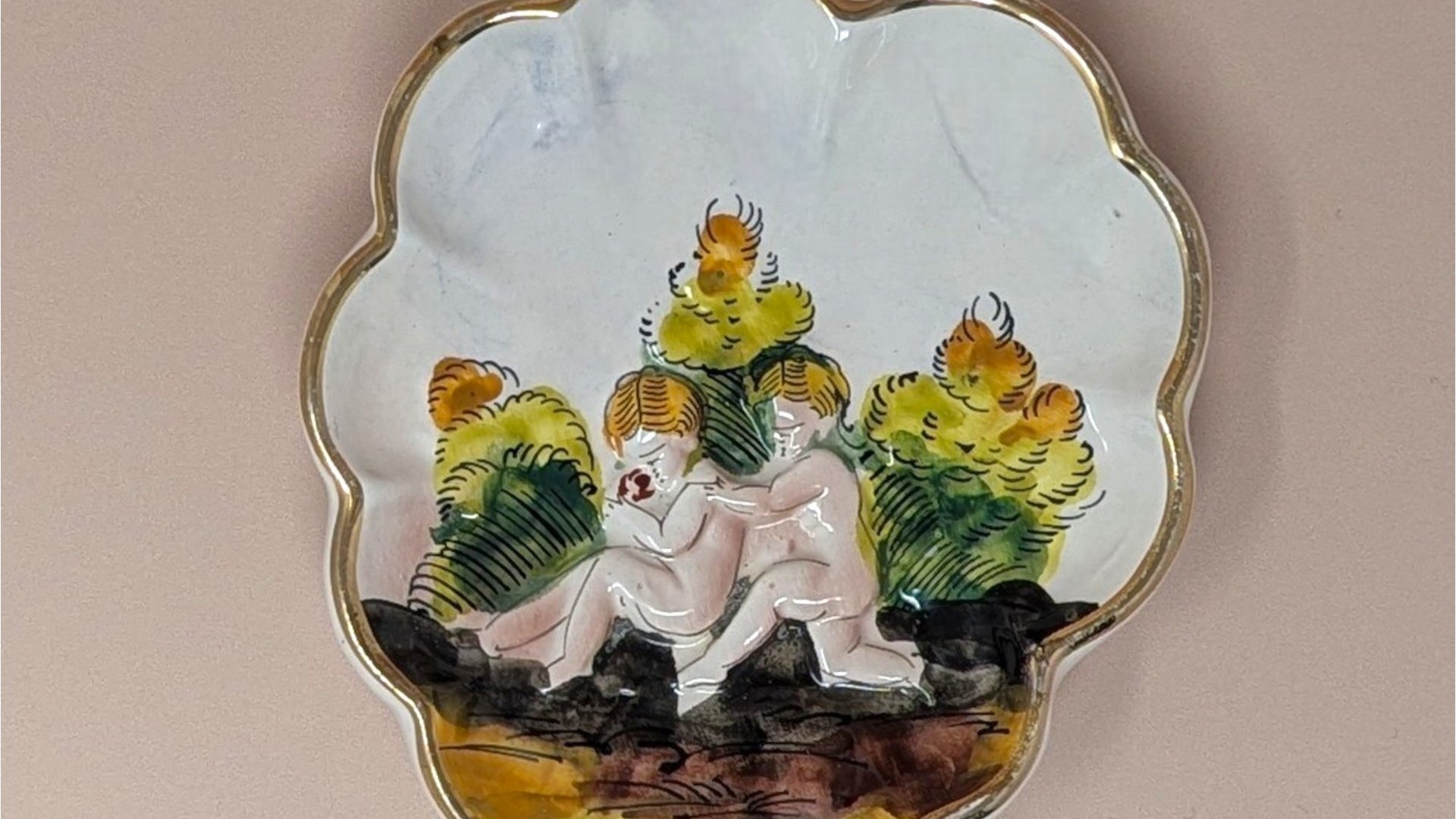 Scalloped Capodimonte porcelain trinket dish with a high-relief embossed scene of two children in a garden. Hand-painted in earthy tones with a gold gilt rim. 5.5 inches wide.