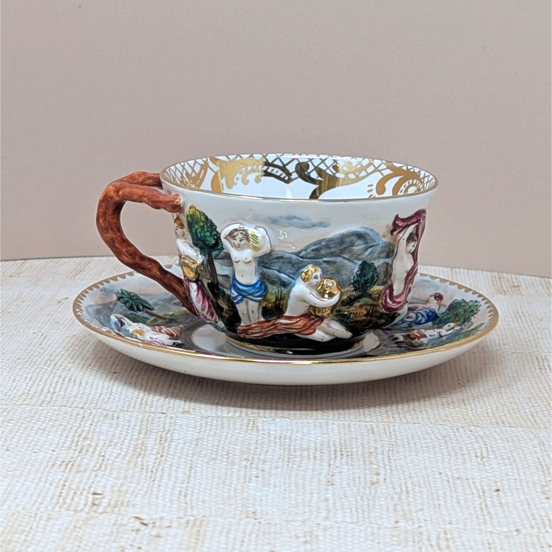 Italian Capodimonte porcelain cup and saucer set. Features high-relief, hand-painted classical figures in a woodland landscape. Intricate gold gilt interior accents and a rustic coral-style handle. Cup 2.5", Saucer 6".