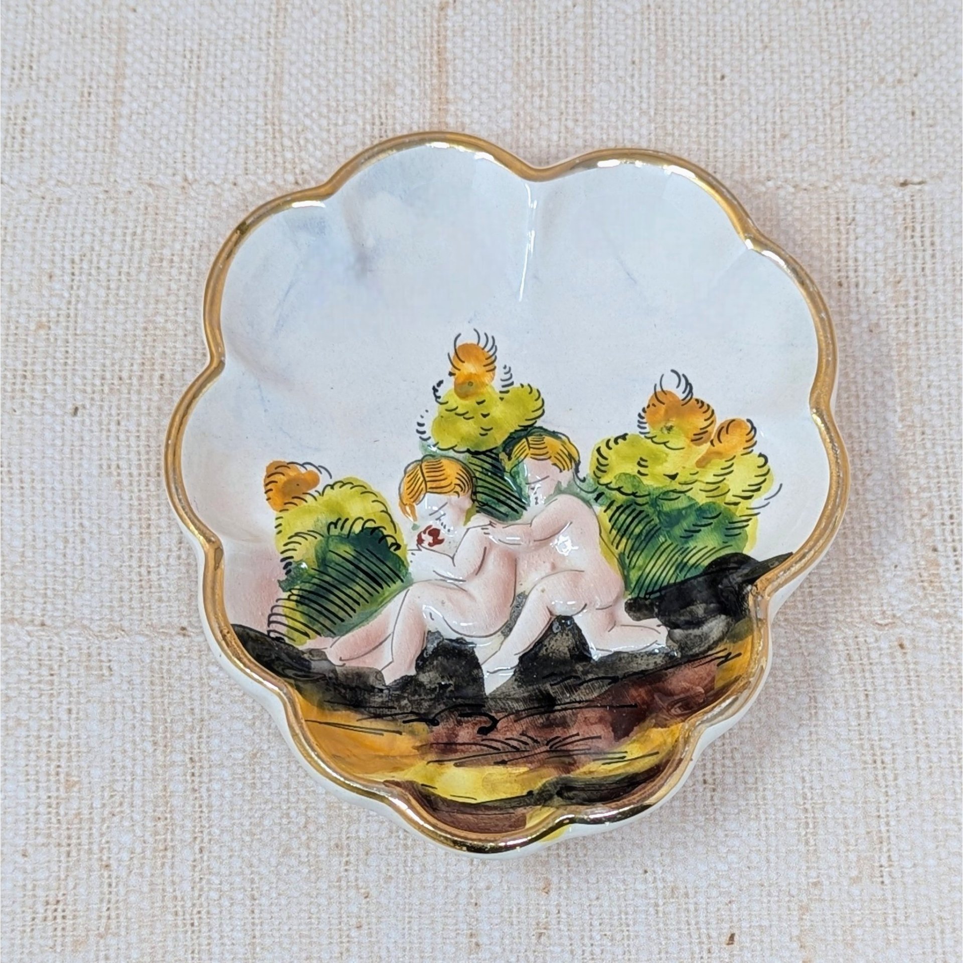Scalloped Capodimonte porcelain trinket dish with a high-relief embossed scene of two children in a garden. Hand-painted in earthy tones with a gold gilt rim. 5.5 inches wide.