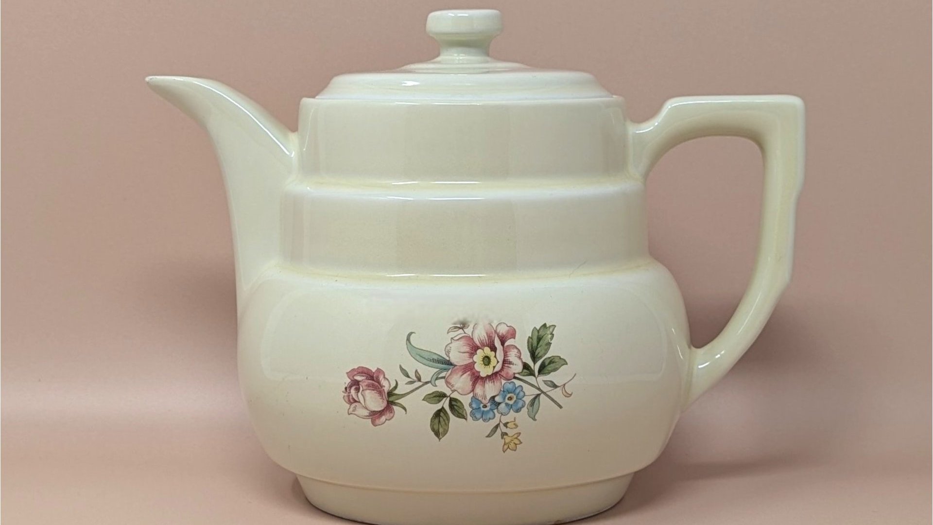 Hall China "Rambling Rose" cream ceramic coffee pot and lid. Features a tiered Art Deco body, an angular handle, and a delicate pink and blue floral spray. 7.75 inches tall.