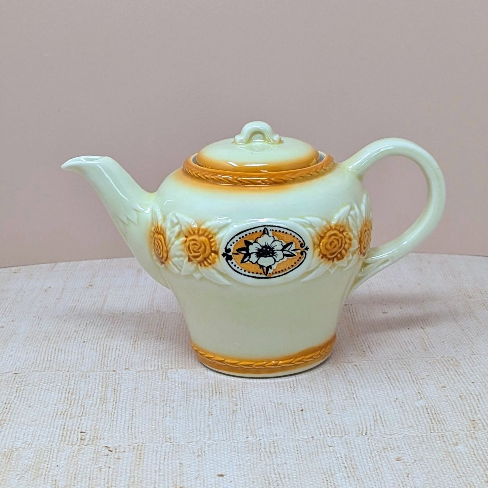 A vintage Porcelier cream-colored teapot with a raised, textured sunflower and rope border in amber-brown tones. A central black and white floral medallion sits within an oval frame. The teapot has a classic rounded shape and matching lid, set against a neutral, warm background.