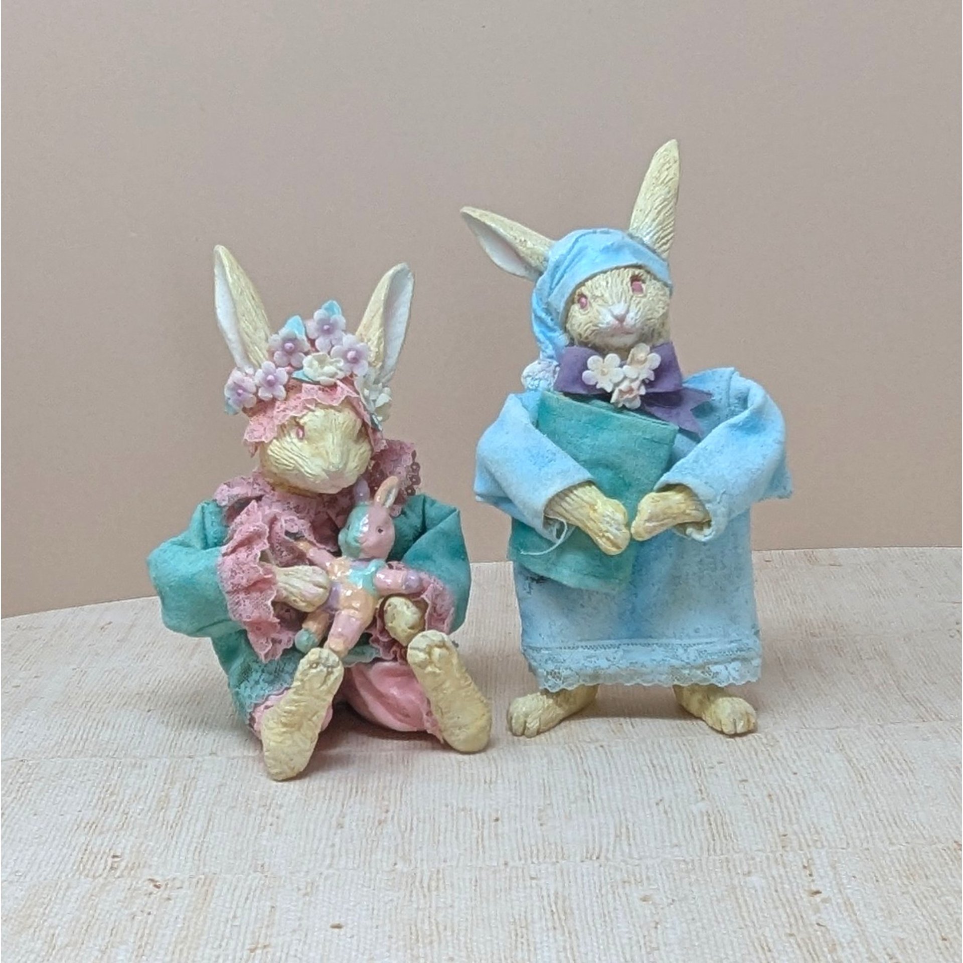 Two whimsical vintage rabbit figurines dressed in pajamas. One wears a blue nightgown and sleeping cap holding a book; the other wears a teal coat and flower crown, holding a small pink rabbit doll.