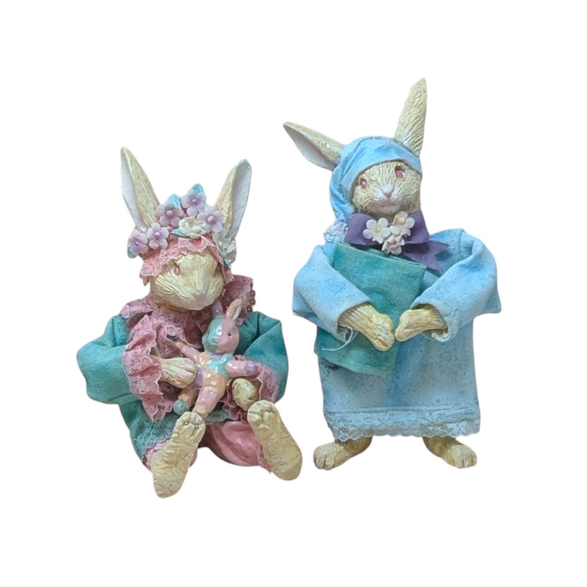 Two whimsical vintage rabbit figurines dressed in pajamas. One wears a blue nightgown and sleeping cap holding a book; the other wears a teal coat and flower crown, holding a small pink rabbit doll.