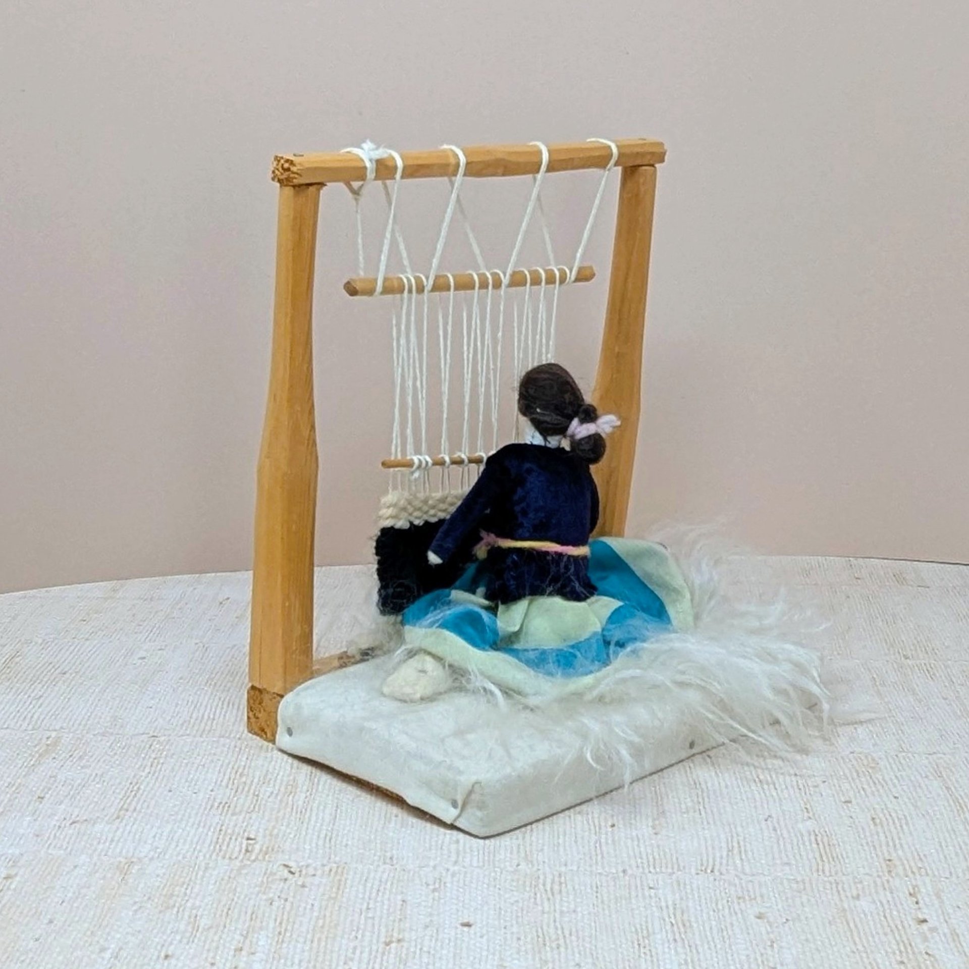 Vintage Navajo weaver doll sitting at a wooden loom. Features a miniature hand-woven textile in progress, a velvet-style bodice, and a turquoise-striped skirt on a faux-fur base. 8 inches tall.