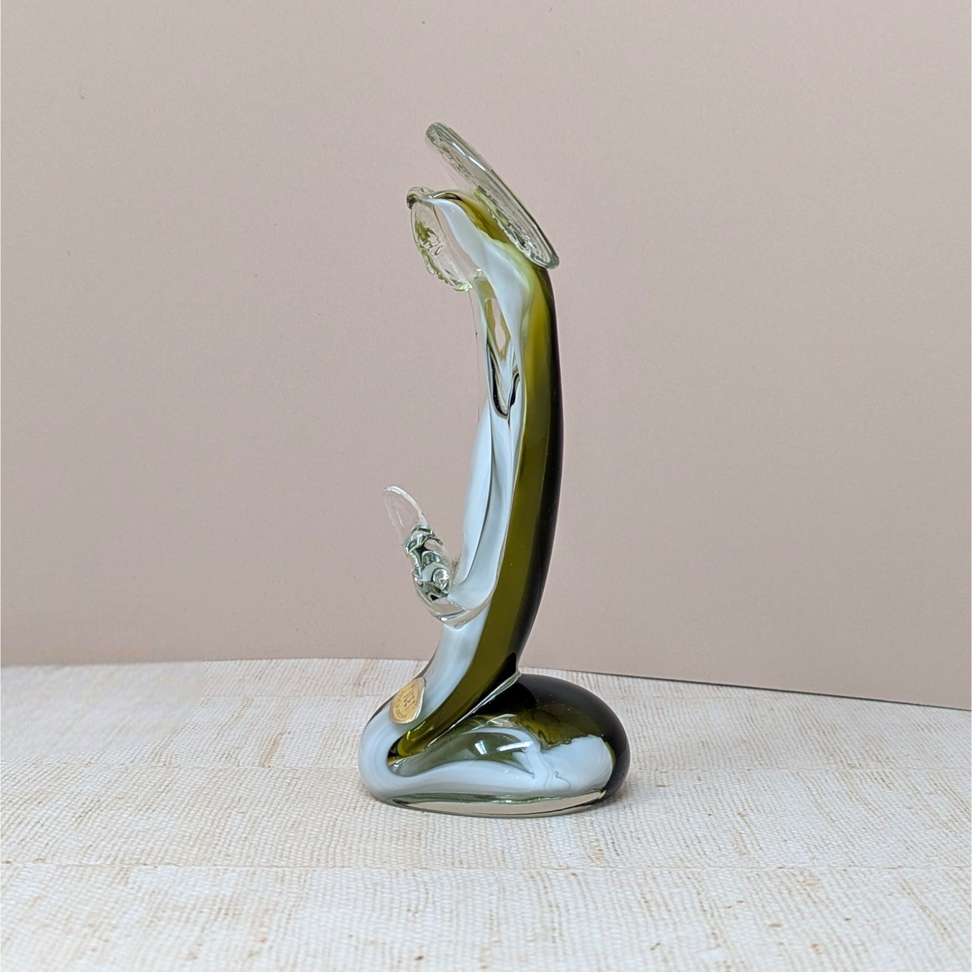A handmade art glass figurine with a smooth, fluid silhouette. The piece features a vibrant emerald green core encased in a clear, glossy glass exterior. Its organic, elongated form tapers elegantly, reflecting light across its polished surface.