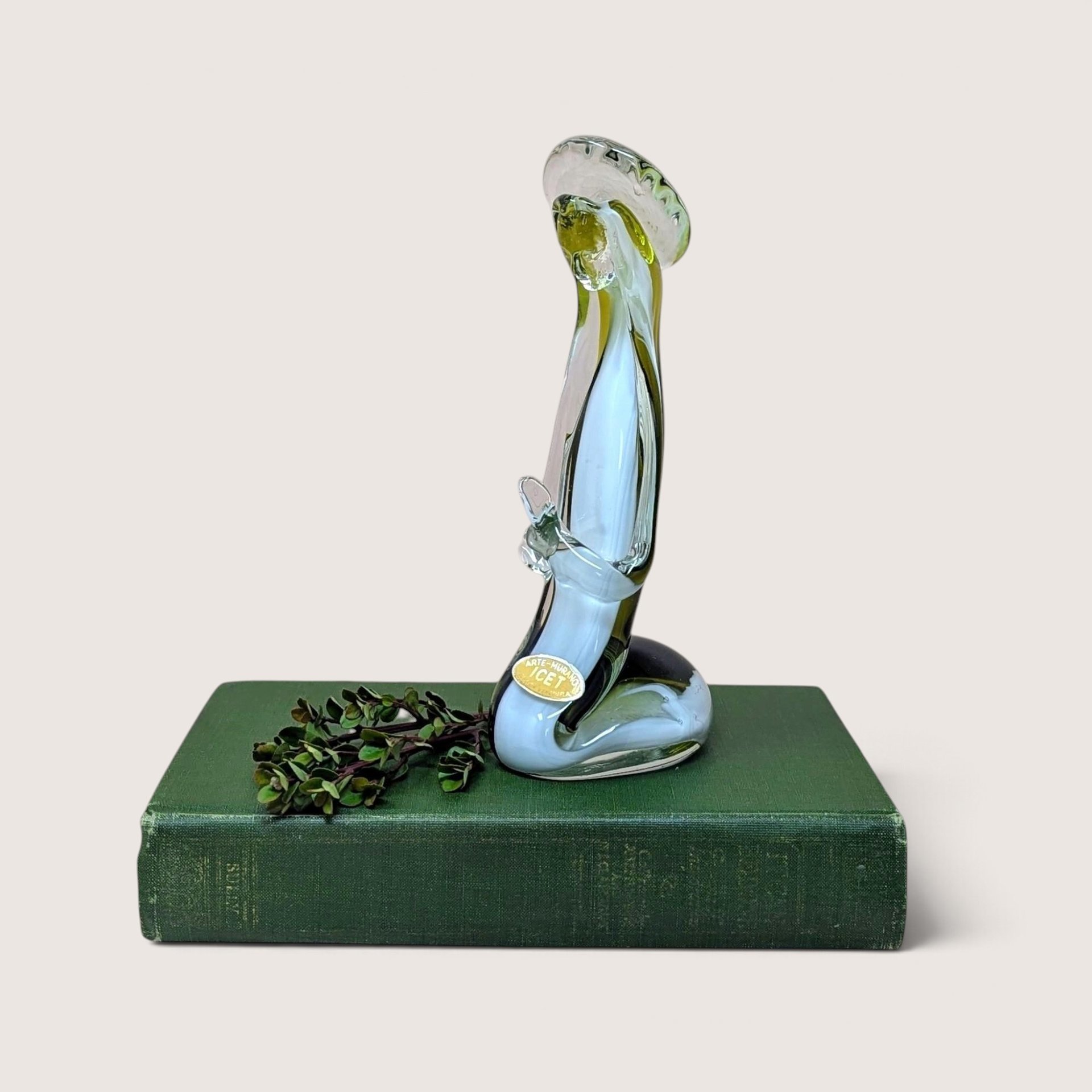 A vertical art glass sculpture of a person in a prayerful pose. A vibrant green inner layer is visible through the thick clear glass exterior, which curves into a stable, weighted base.