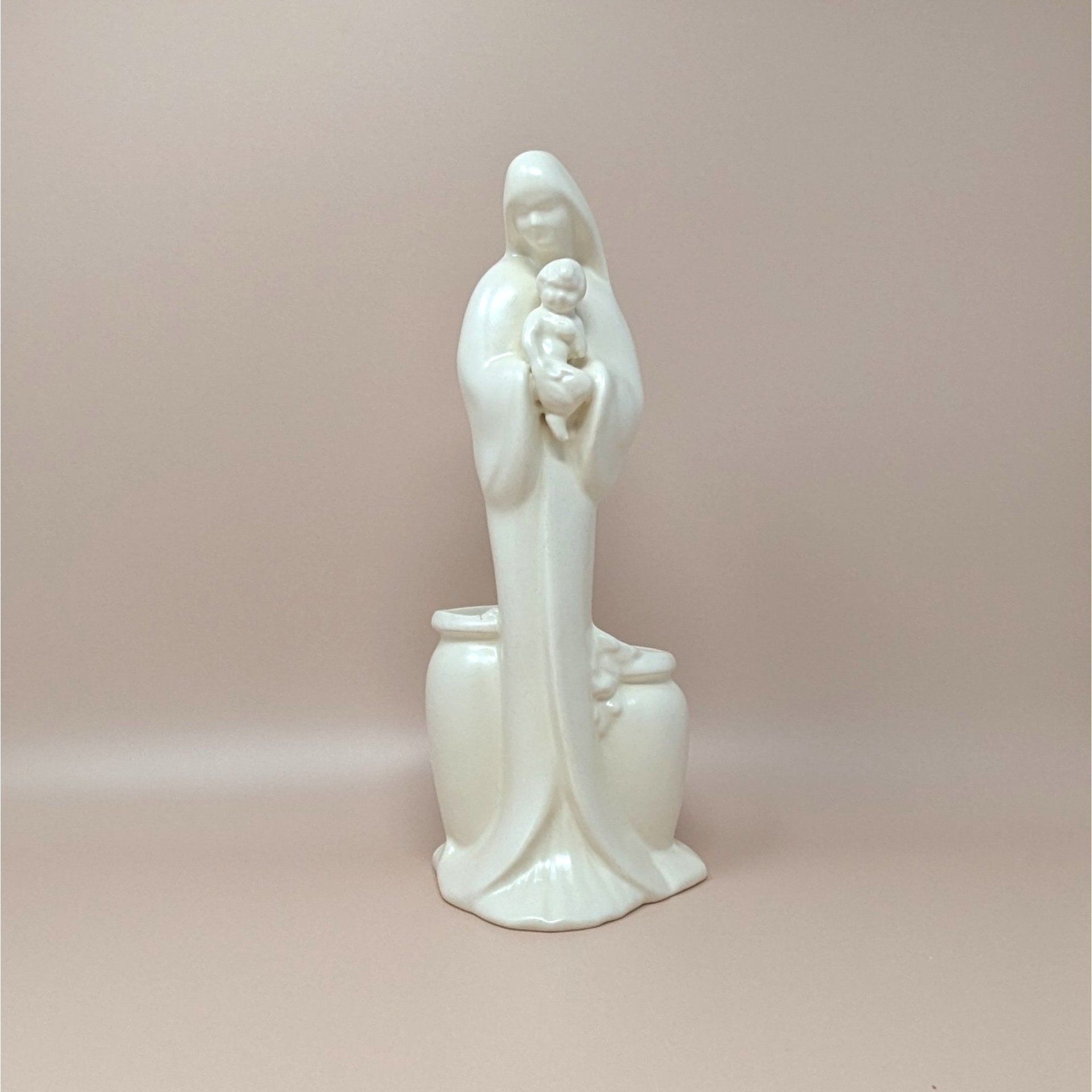 Royal Haeger white pottery planter featuring a minimalist Madonna holding the Christ Child. High-gloss white glaze with two small planter basins at the base. 11.75 inches tall.A 11.75-inch tall cream-colored semi-gloss ceramic figurine of Mary holding the infant Jesus, with two integrated vase-style planter pockets at the base.