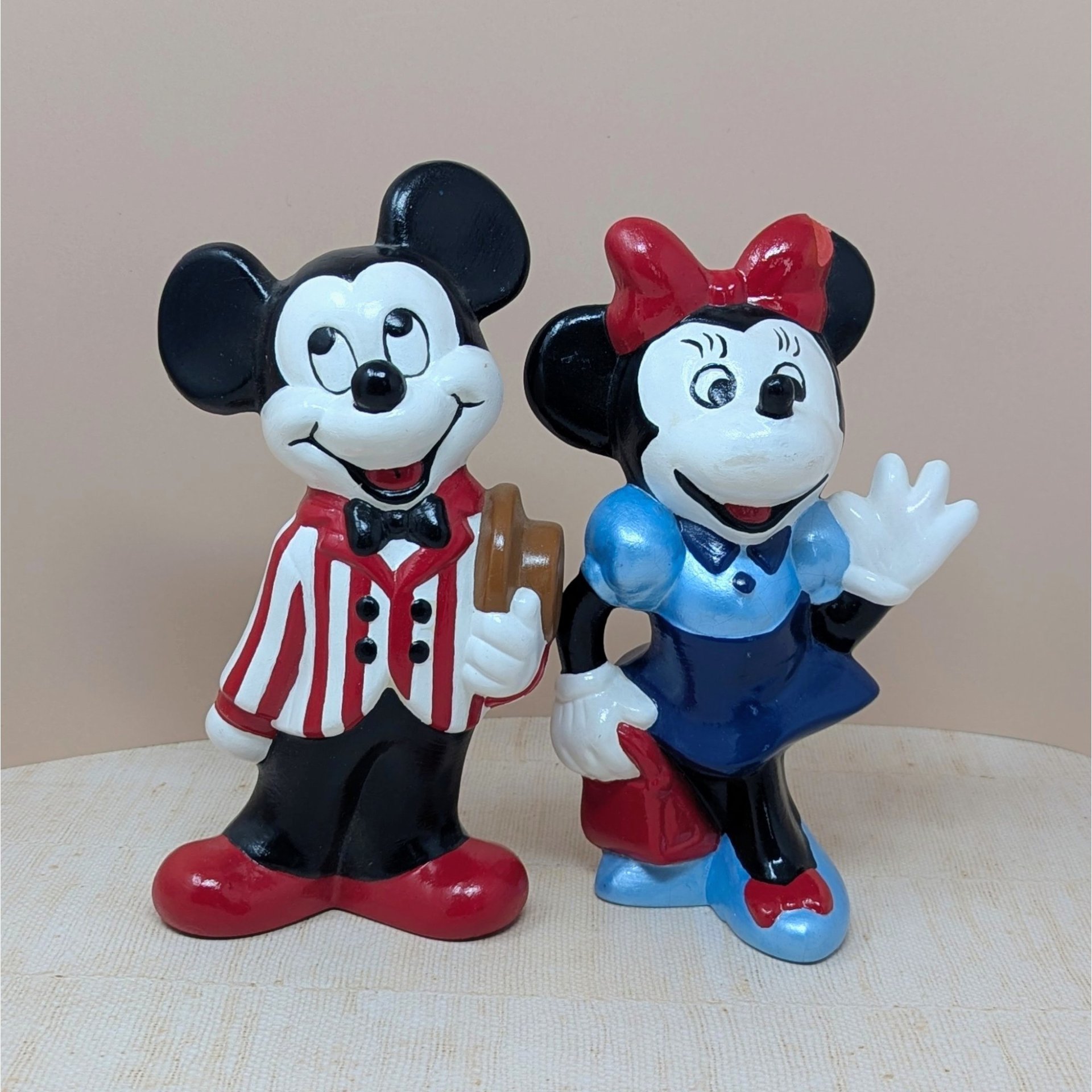 Set of two vintage 9-inch hand-painted Disney figurines. Dapper Mickey in a red-and-white striped jacket holding a brown hat. Minnie in a blue dress and red bow waving. Ceramic style.