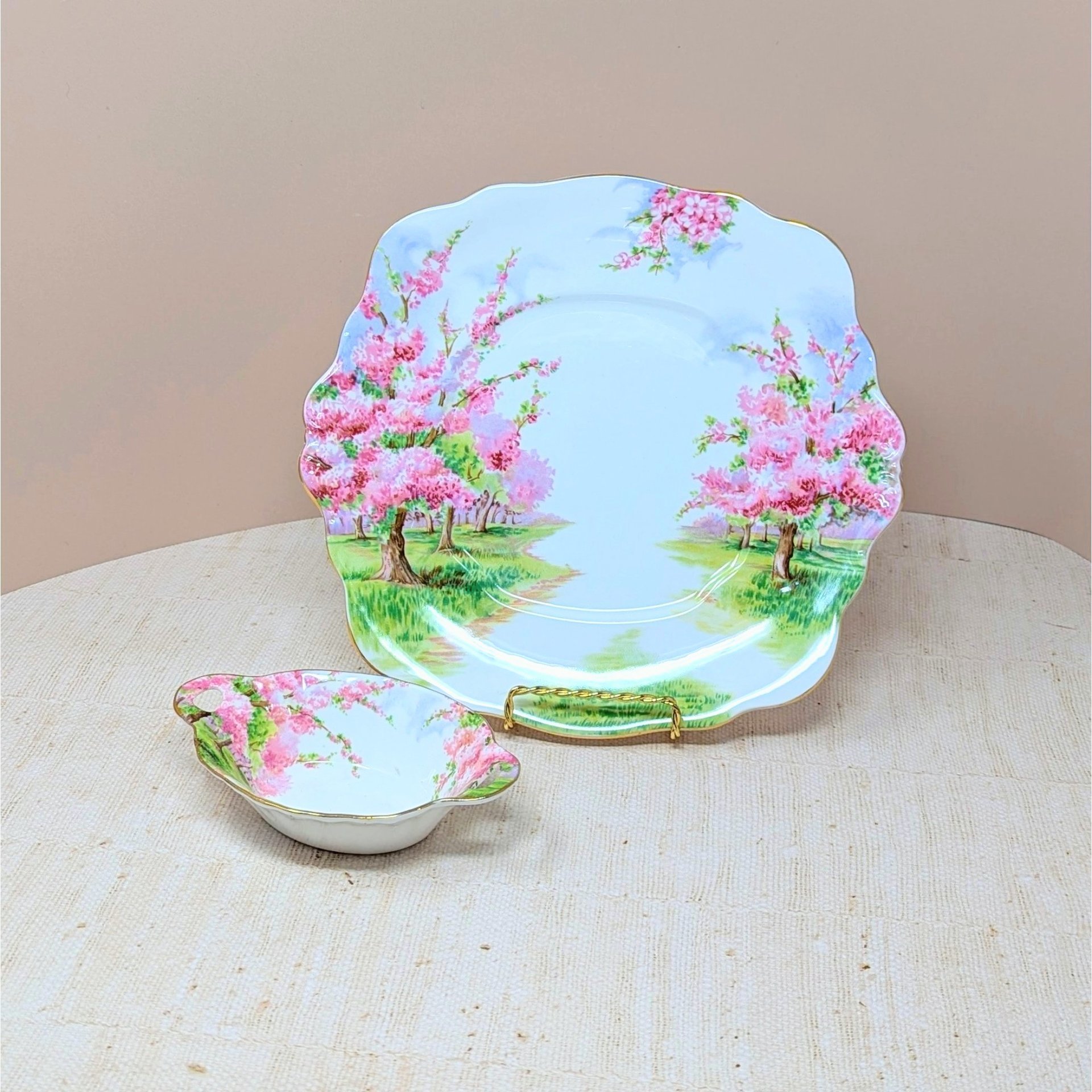 Two-piece Royal Albert Blossom Time bone china set. Includes a 9.75-inch square handled cake plate and a 5.5-inch oval handled sweet meat dish. Features pink blossom trees and gold-trimmed scalloped edges.