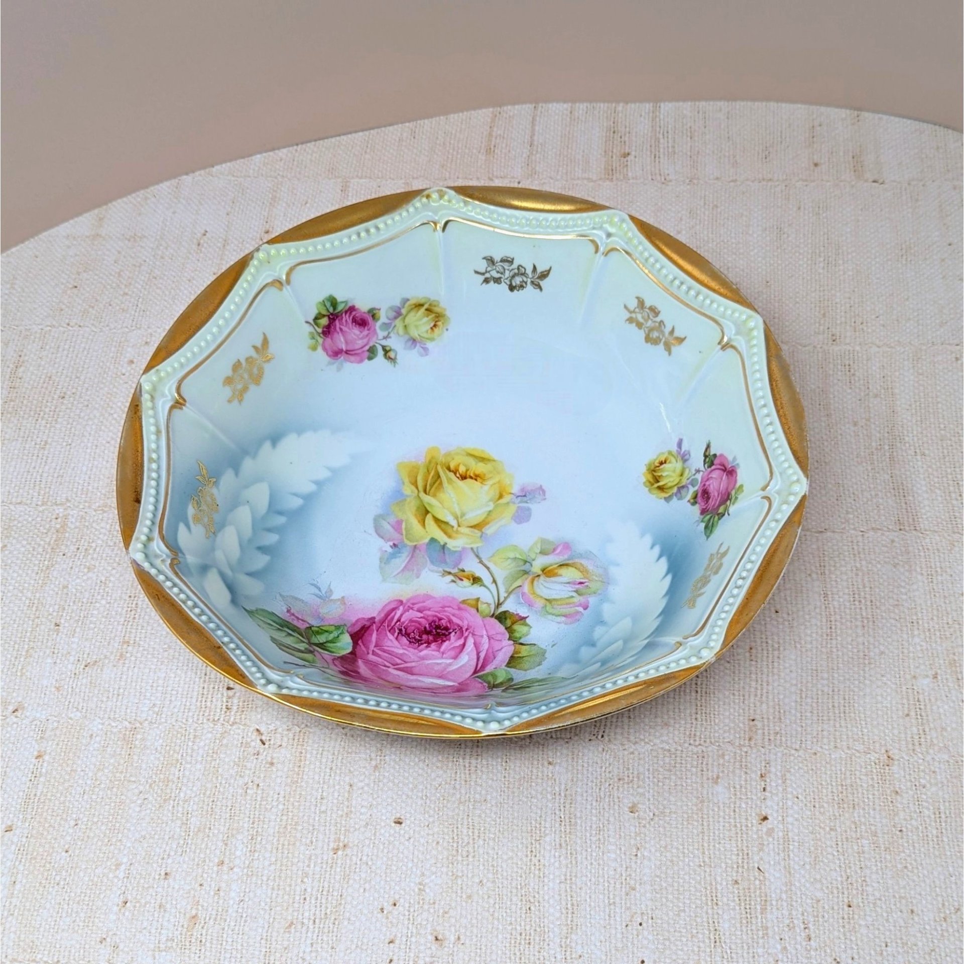 Scalloped octagonal porcelain bowl. Pale blue ombre interior with large pink and yellow roses. Wide rim features heavy gold gilt with a delicate beaded relief texture.