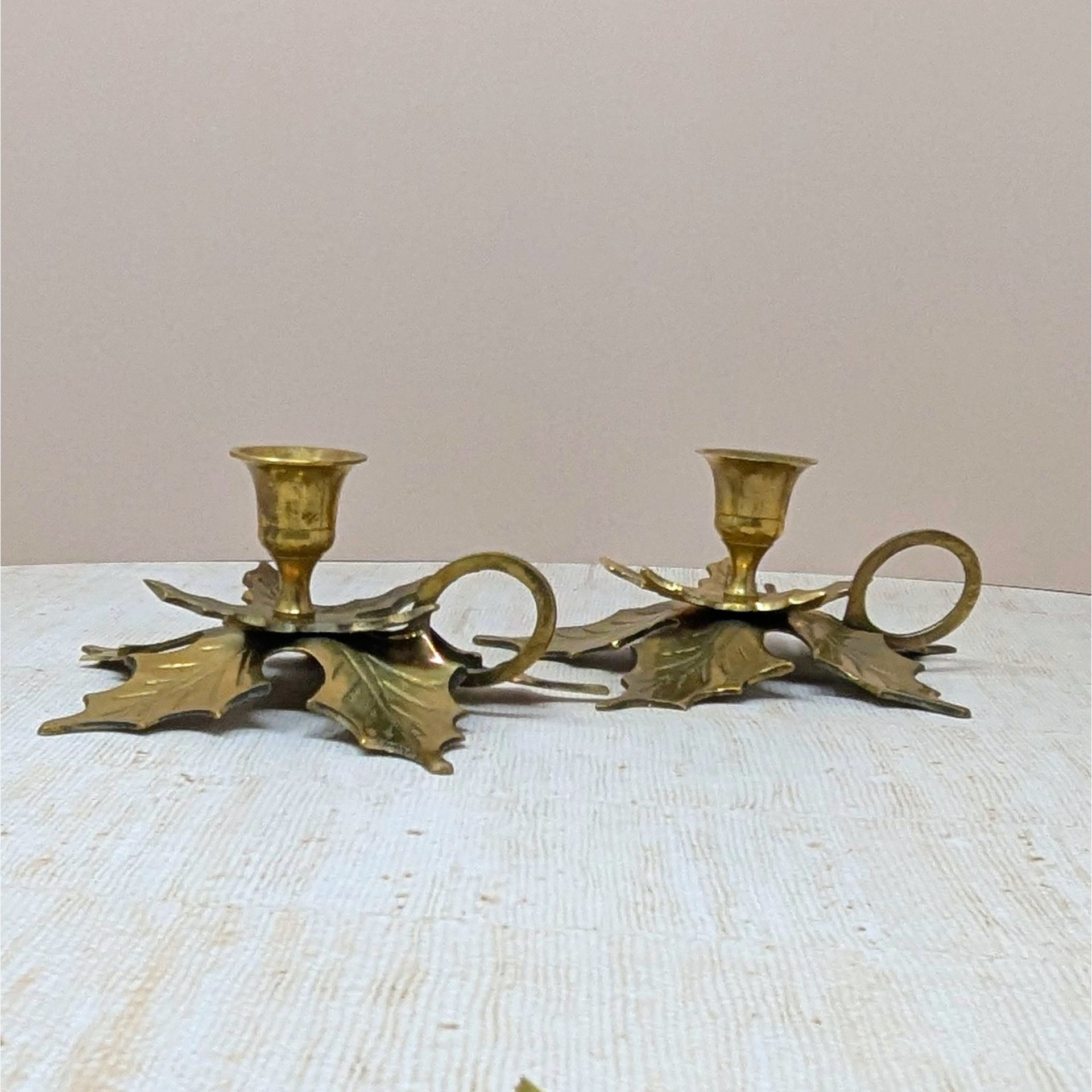 A pair of solid brass chamberstick candle holders. Each base is shaped like two large, overlapping maple leaves with detailed veining. Features a finger loop handle and a single tulip-style taper cup.