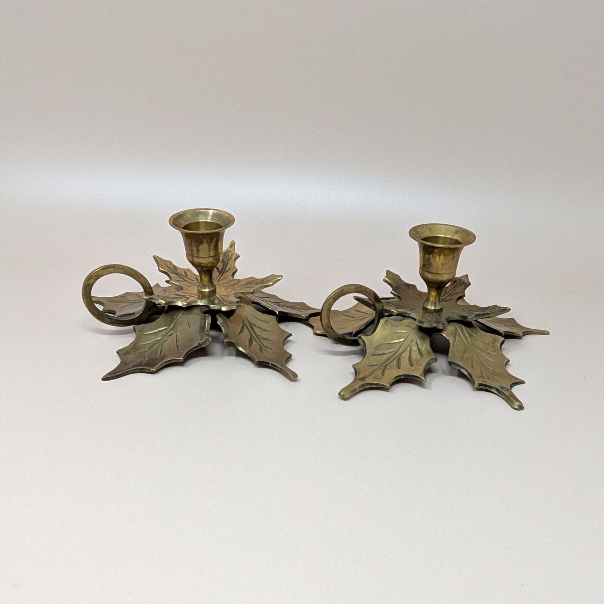 A pair of solid brass chamberstick candle holders. Each base is shaped like two large, overlapping maple leaves with detailed veining. Features a finger loop handle and a single tulip-style taper cup.