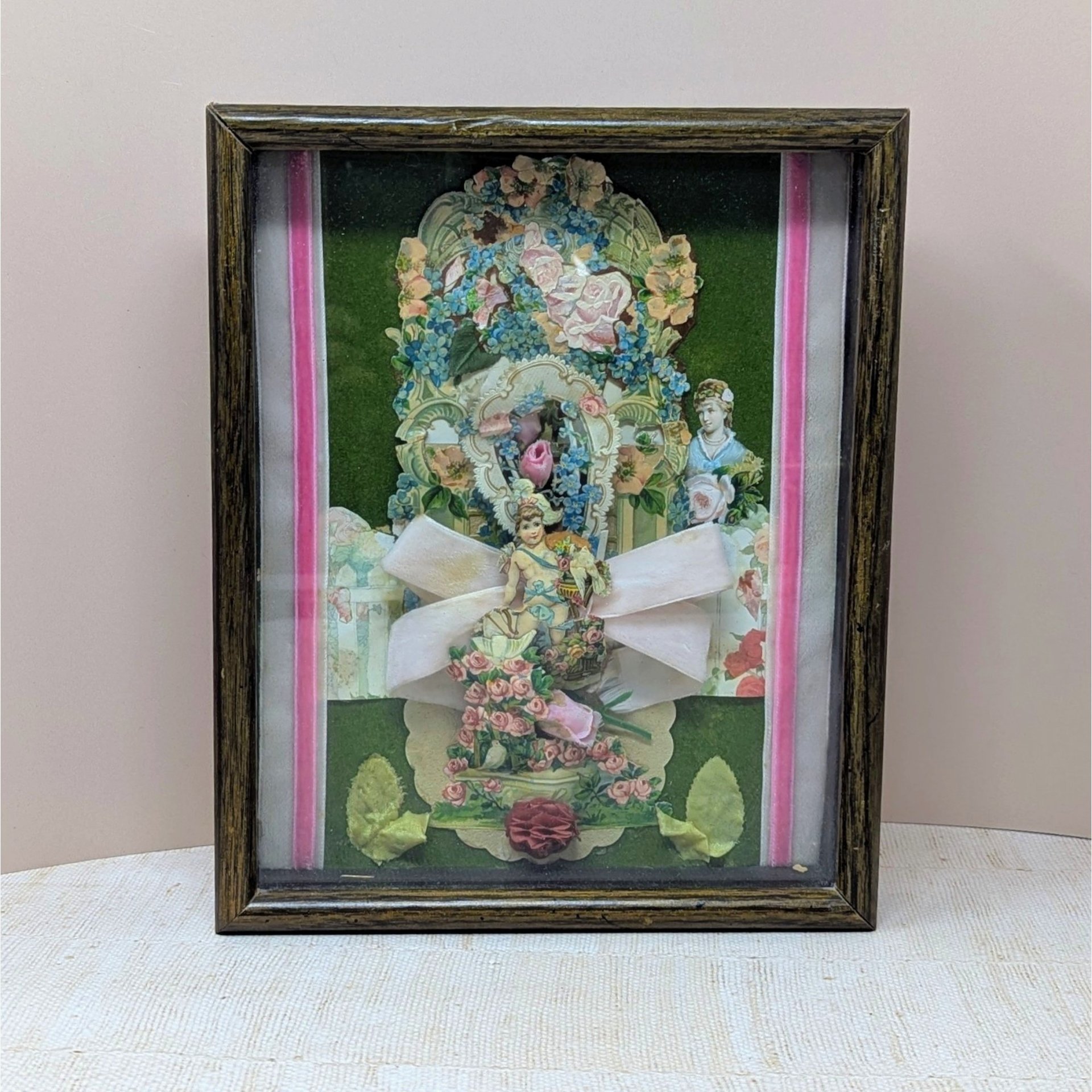 A vertical shadow box frame containing a Victorian-style paper collage. Features a cherub, floral lithographs, and a large pink velvet bow against a forest green velvet background. Housed in a dark wood-grain frame.