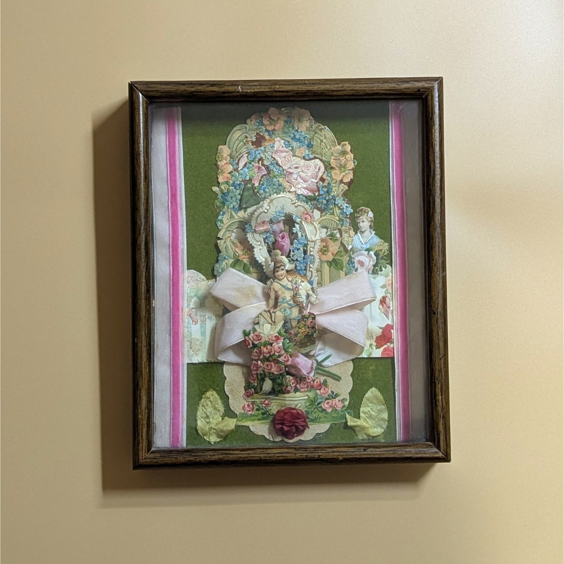 A vertical shadow box frame containing a Victorian-style paper collage. Features a cherub, floral lithographs, and a large pink velvet bow against a forest green velvet background. Housed in a dark wood-grain frame.