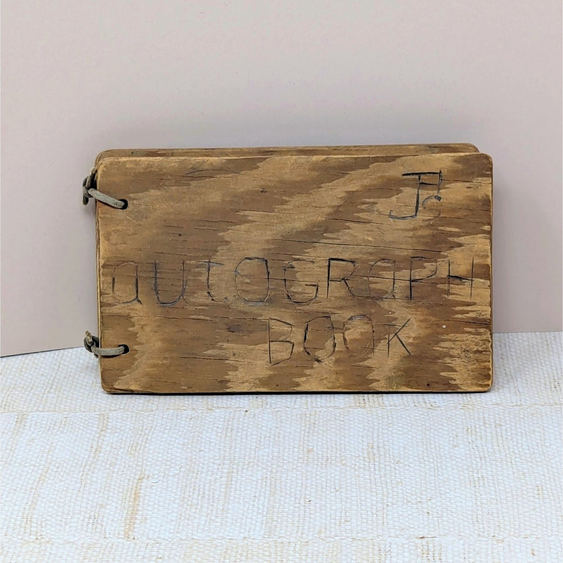 A small, rectangular autograph book with a natural wood grain cover. The words "Autograph Book" are hand-carved into the wood. Two metal rings bind the pages on the left. Rustic, weathered texture with a warm honey-oak finish.
