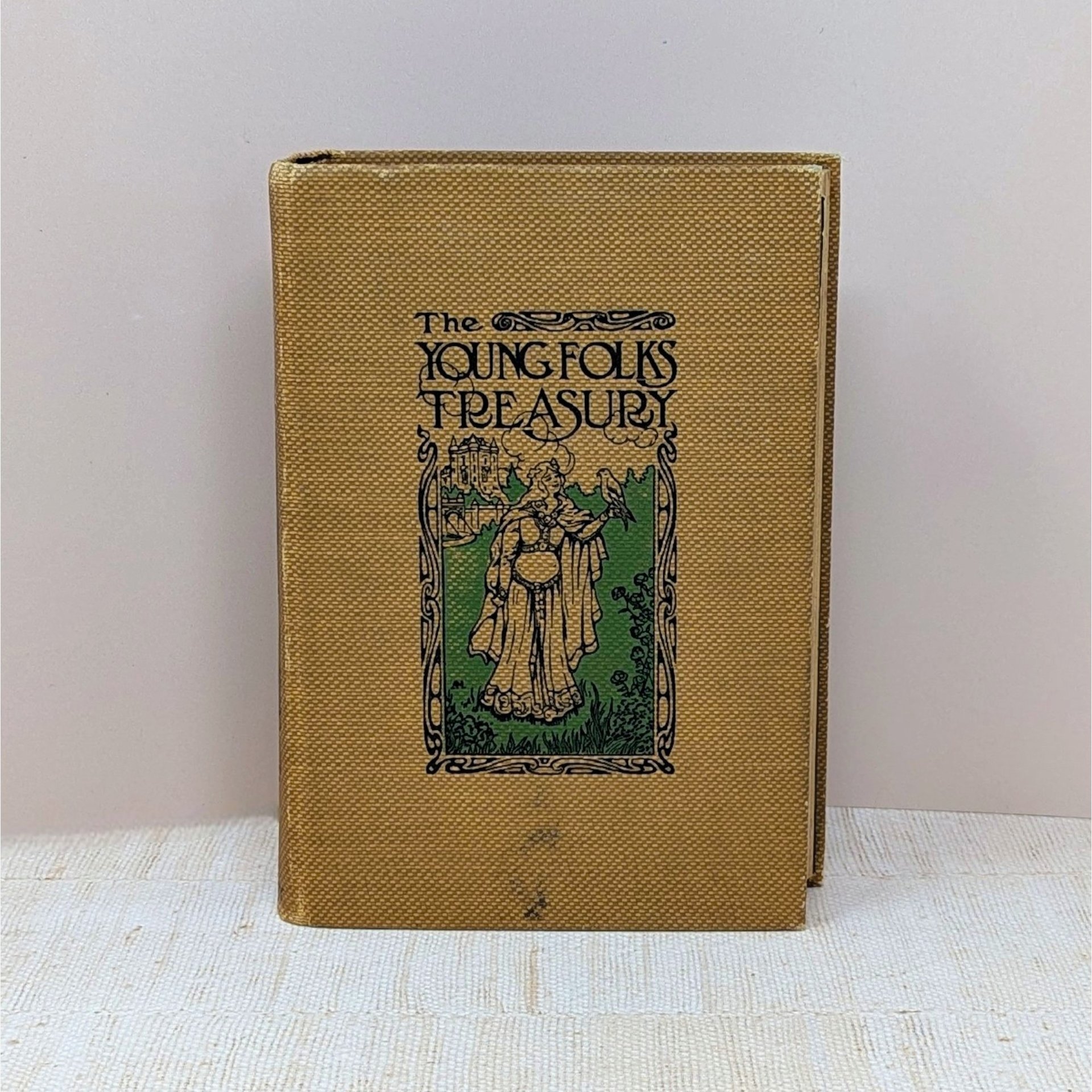 Antique book with a golden-yellow textured cloth cover. Features an Art Nouveau illustration of a woman with a hawk and a castle. Titled "The Young Folks Treasury" in stylized black font. Aged, tactile fabric finish.