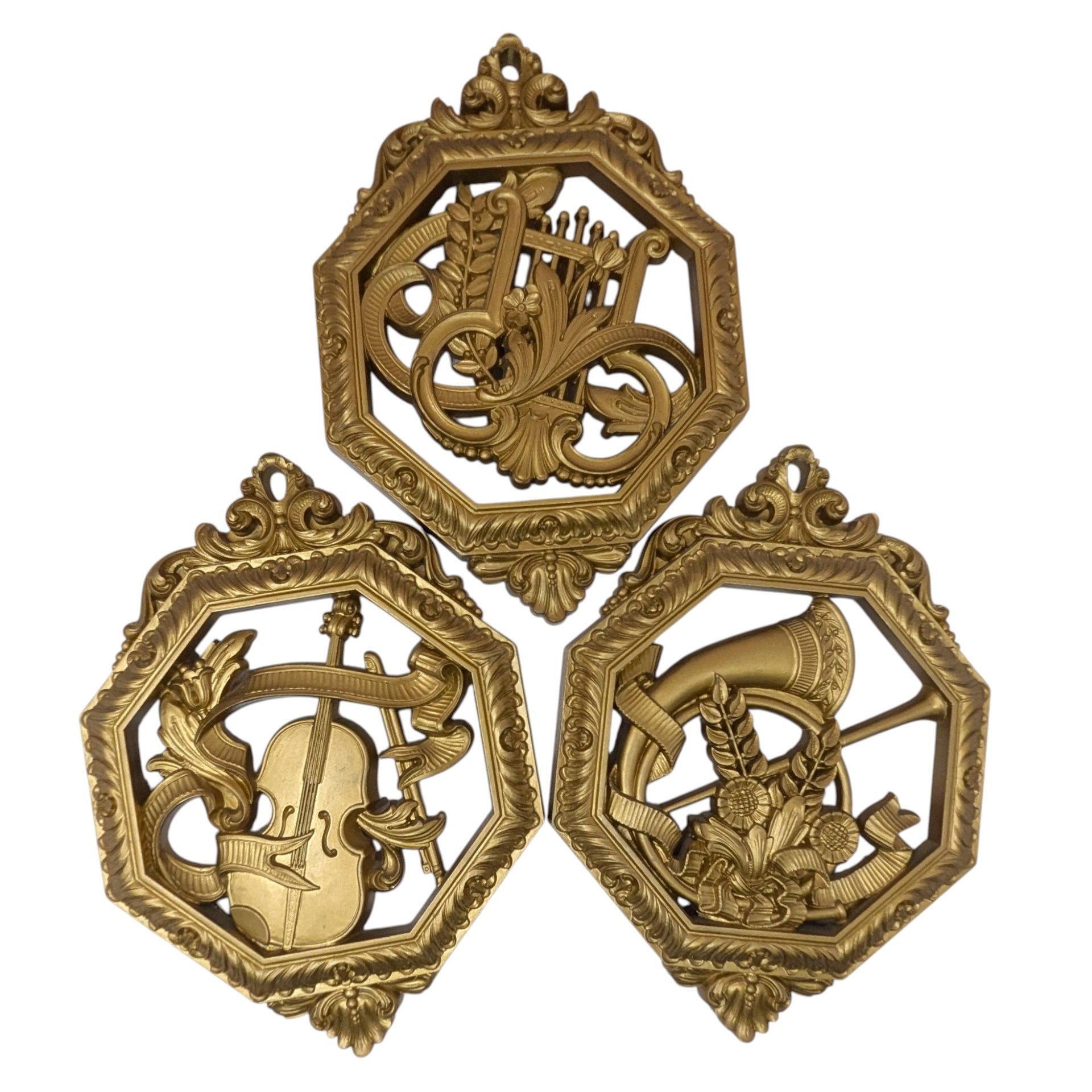 Set of three octagonal gold-toned wall plaques. Each features a different relief-style musical instrument: a lyre, a horn, and a violin. Ornate open-work frames with a metallic gilt finish.