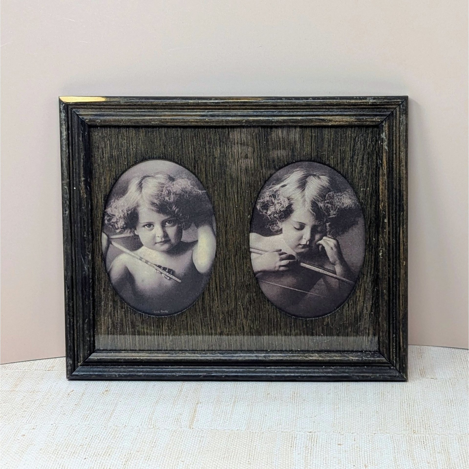 A dark wood frame with two oval cutouts. Inside are sepia-toned prints of a young child with curly hair and wings, depicting "Angel Awake" looking at the viewer and "Angel Asleep" with eyes closed. Rustic wood grain texture.