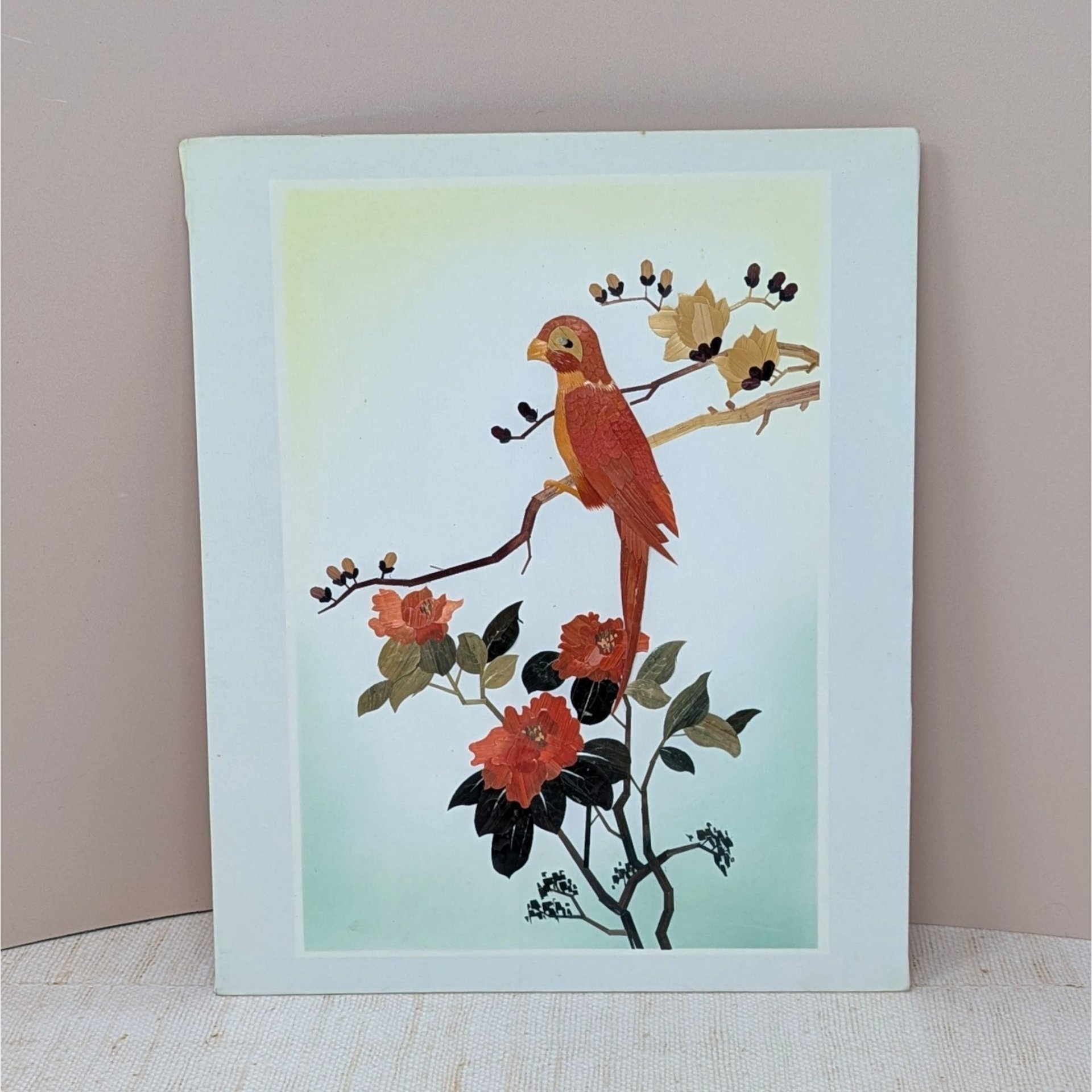 A vertical rectangular wall art piece featuring a bird perched on a branch, meticulously crafted from layered bamboo. Warm sienna and ochre tones are accented by deep red floral blooms. Framed with a clean white and pink double mat.
