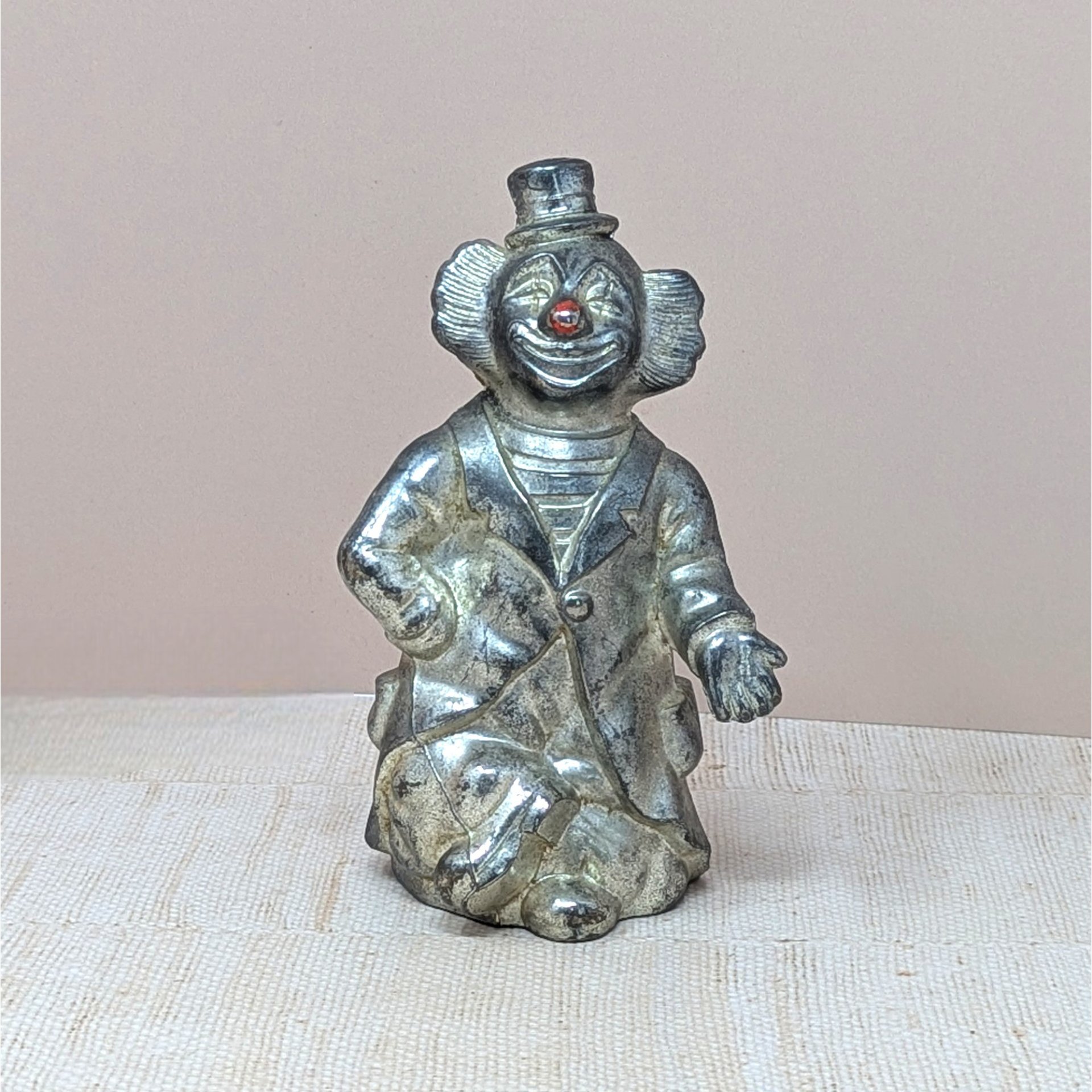 A vertical silver-toned metal bank shaped like a circus clown in a top hat and suit. Features an aged metallic patina with a small red detail on the nose. Sculptural, heavy-duty texture with a slot on the reverse.