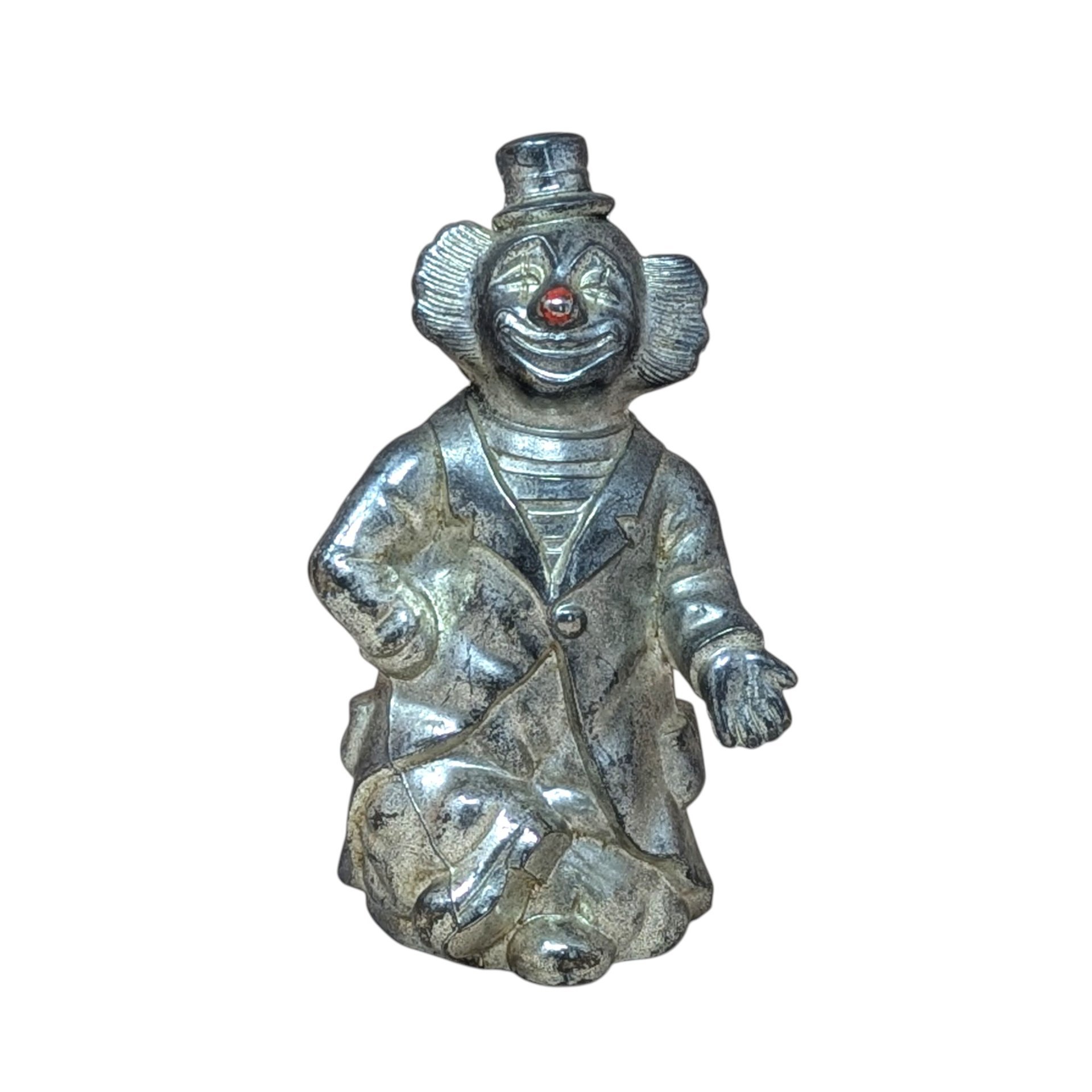 A vertical silver-toned metal bank shaped like a circus clown in a top hat and suit. Features an aged metallic patina with a small red detail on the nose. Sculptural, heavy-duty texture with a slot on the reverse.