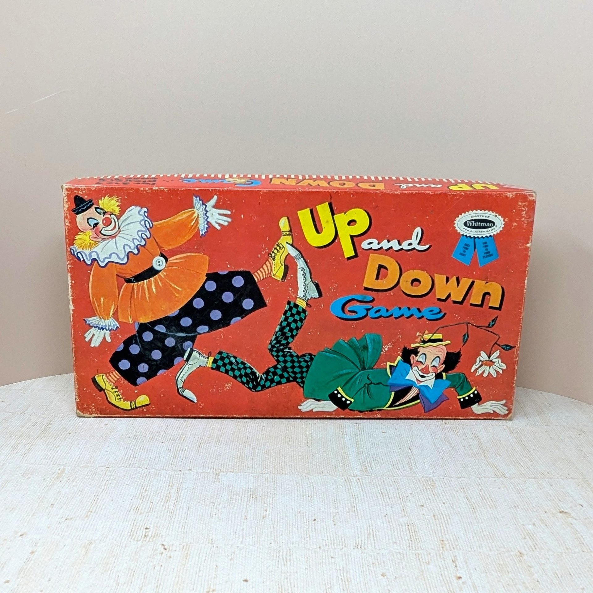 A horizontal vintage board game box for "Up and Down Game" by Whitman. Features two whimsical clowns in colorful costumes against a vibrant red background. Matte paper texture with visible edge wear and aged patina.