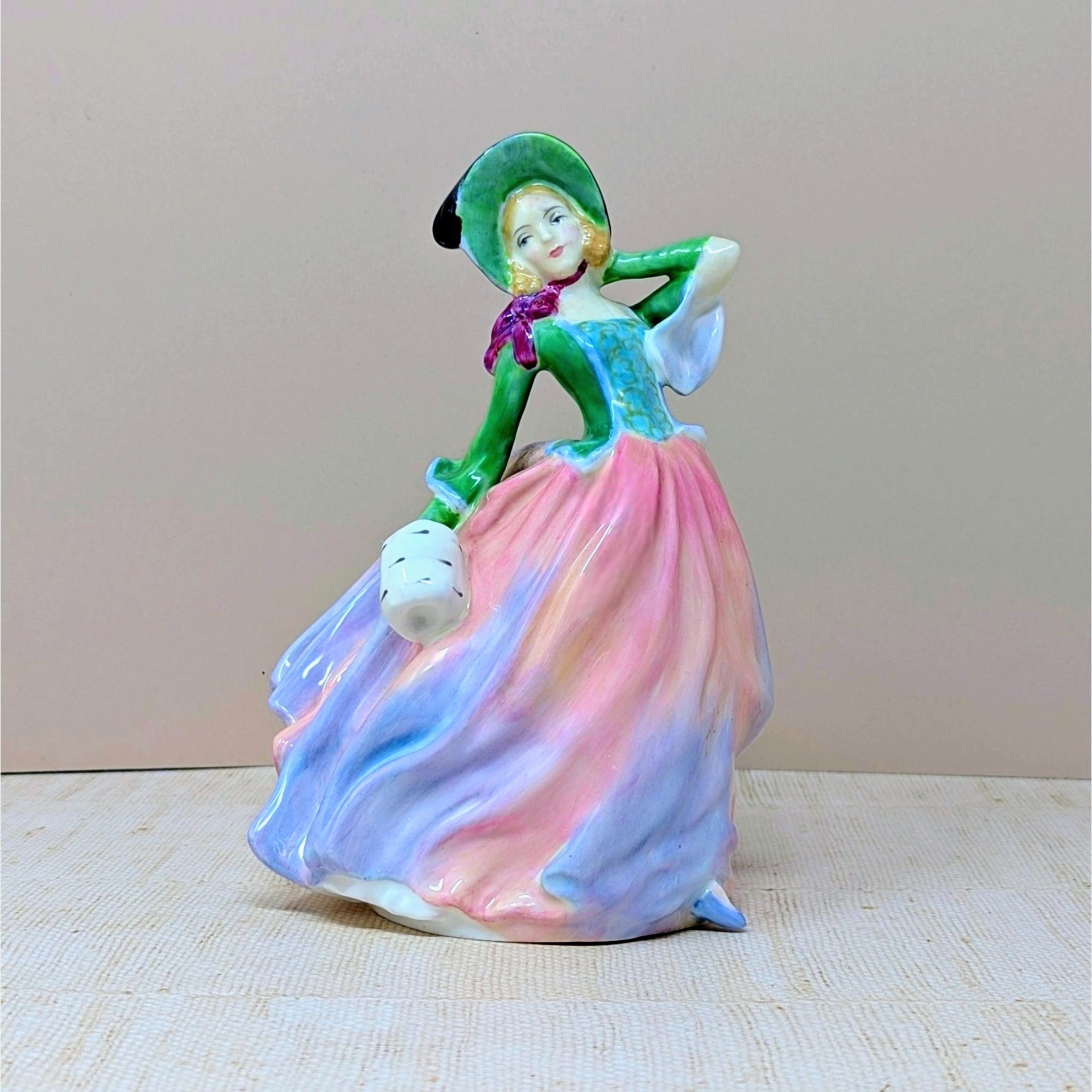 A vertical porcelain figurine of a woman in a wind-swept gown. She wears a vibrant green bodice, a wide-brimmed hat, and a voluminous skirt in blended shades of pink, peach, and lavender. Glossy, fine-bone china texture.
