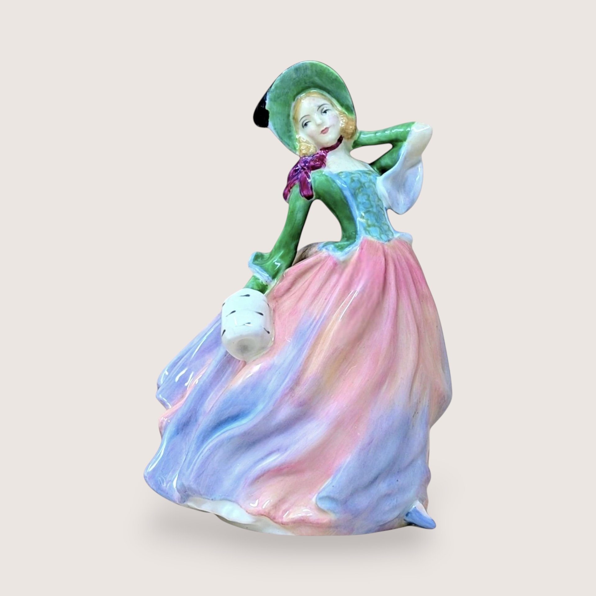 A vertical porcelain figurine of a woman in a wind-swept gown. She wears a vibrant green bodice, a wide-brimmed hat, and a voluminous skirt in blended shades of pink, peach, and lavender. Glossy, fine-bone china texture.