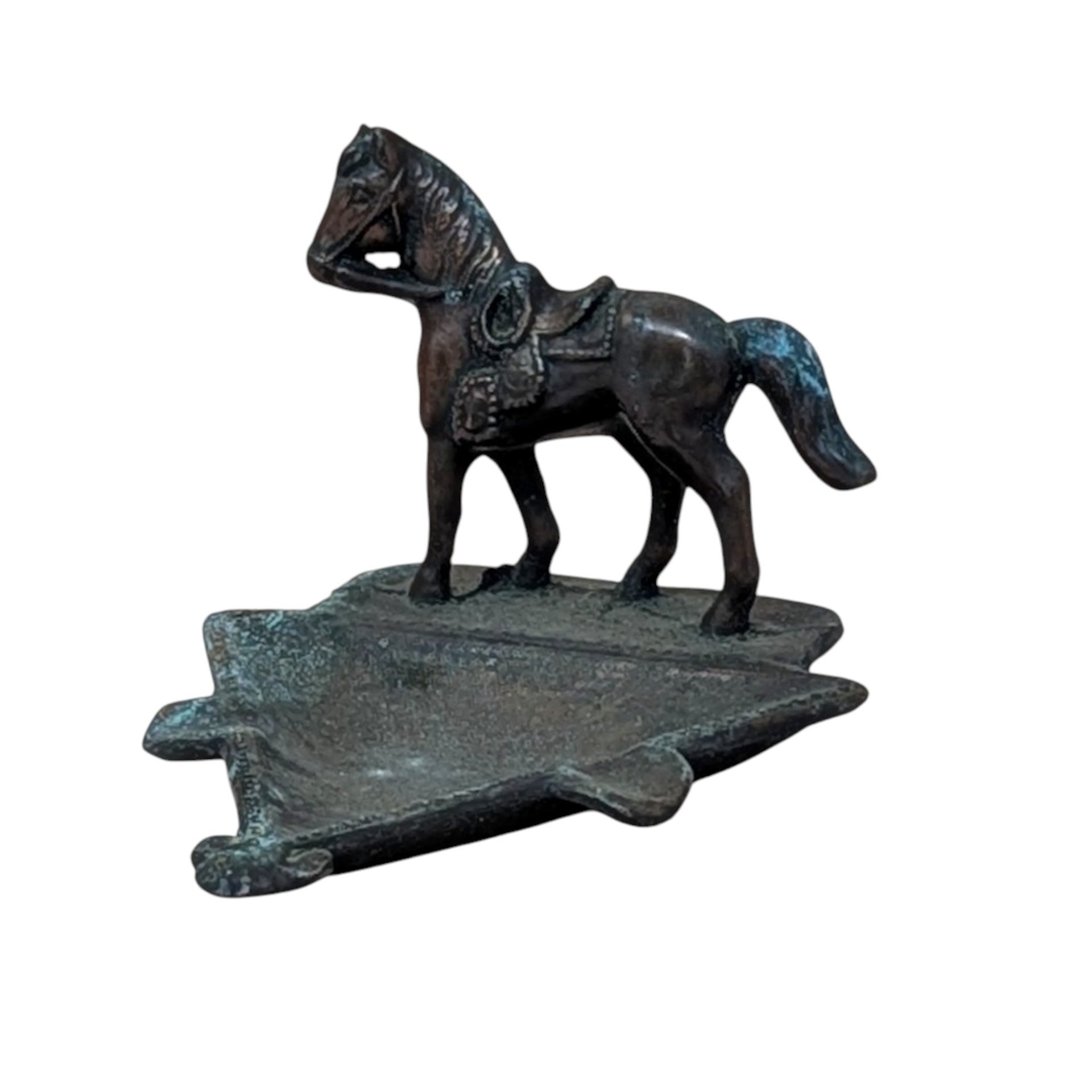 A vertical cast metal ashtray featuring a saddled horse figurine standing on a triangular base. The metal has a dark bronze patina with heavy green verdigris oxidation throughout. Sturdy, industrial texture with period-correct relief details.
