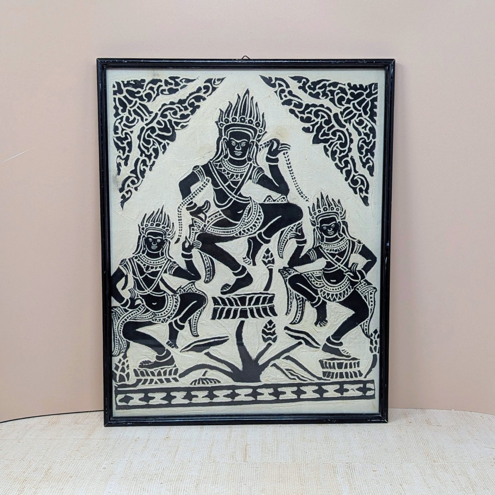 A vertical framed art piece on textured rice paper featuring three Apsara dancers in traditional Cambodian headdresses and poses. Bold black ink on a cream-white background. Framed in a minimalist black wood frame.